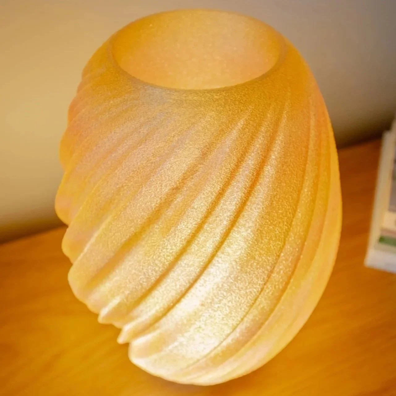 	
A glowing, oval yellow 3d printed lamp with a textured surface, sitting on a modern white tripod base on a wooden table, with a potted plant in a white textured pot in the background.