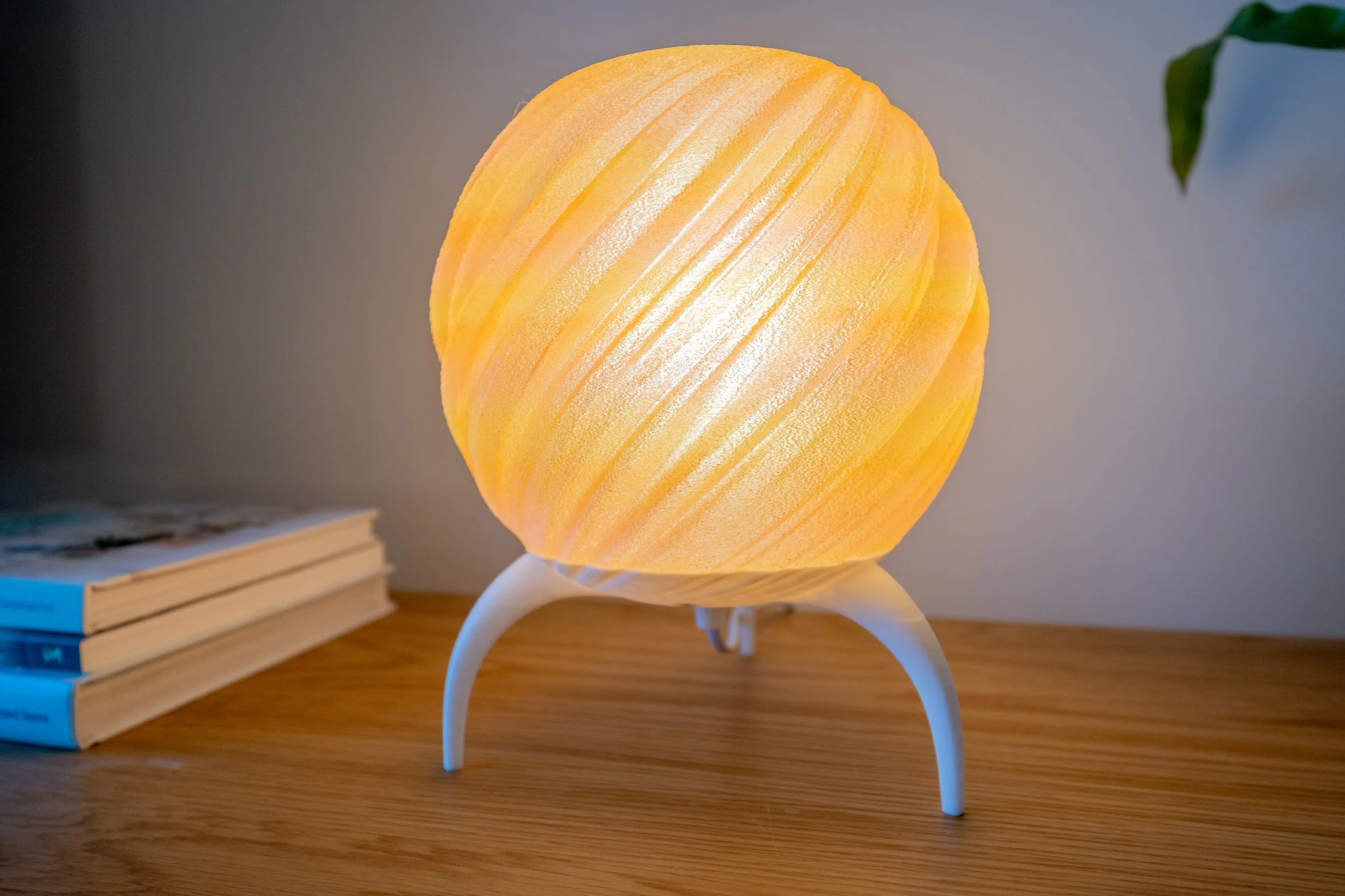 A spherical orange 3d printed lamp with diagonal textured lines, standing on three white curved legs on a wooden surface