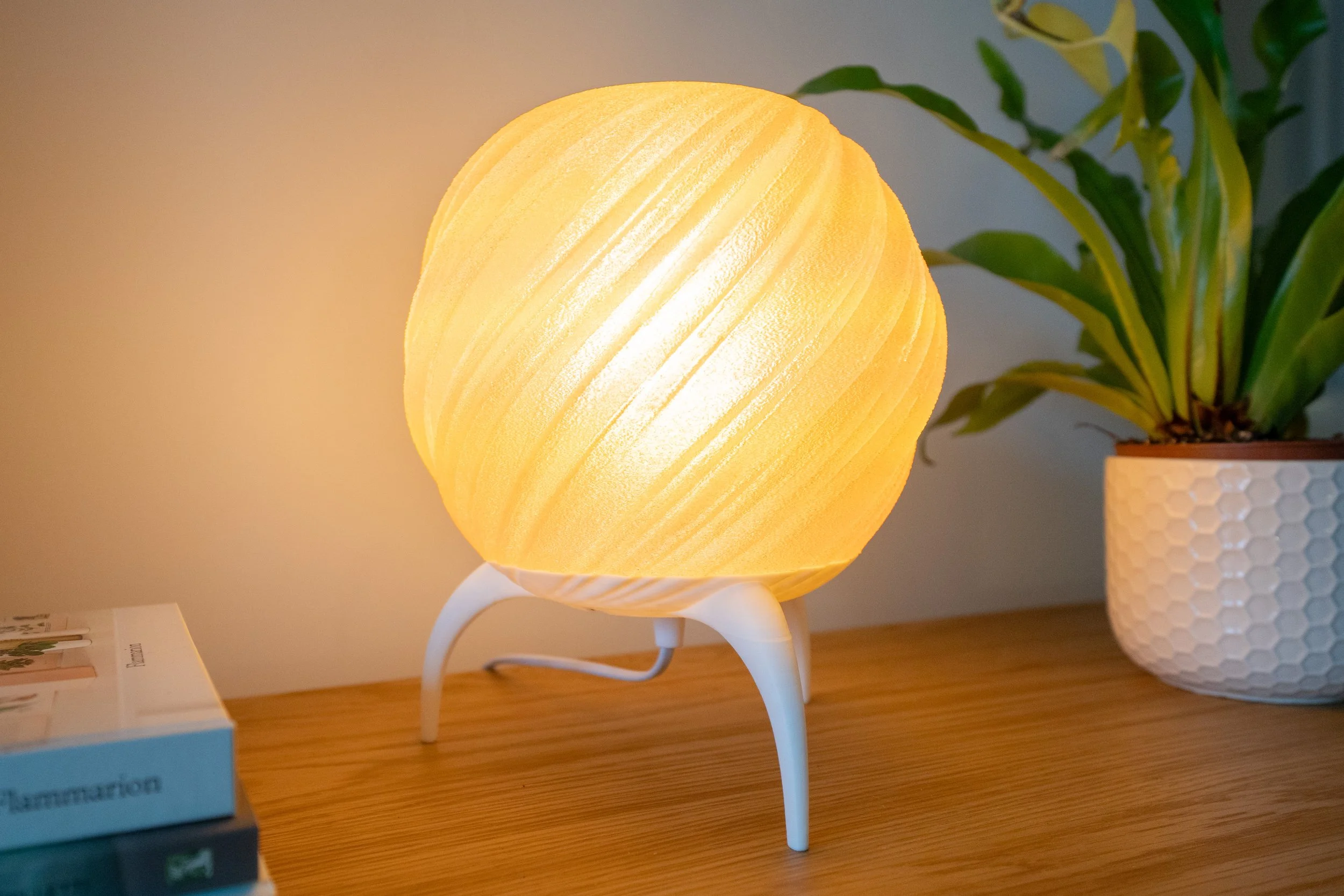 A glowing, spherical yellow 3d printed lamp with a textured surface, sitting on a modern white tripod base on a wooden table, with a potted plant in a white textured pot in the background.