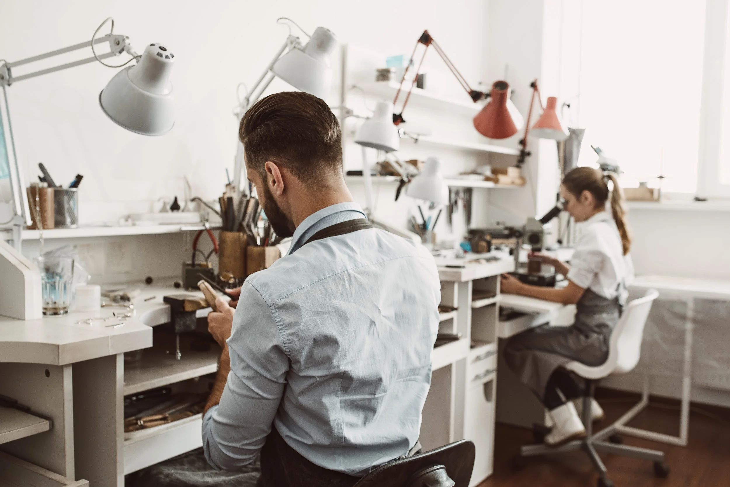 Mann in Arbeitskleidung arbeitet an einem kleinen technischen Gerät, im Hintergrund eine junge Frau, die durch ein Mikroskop schaut, beides in einem hellen, modernen Labor mit weißen Wichts und flexiblen Arbeitslampen.