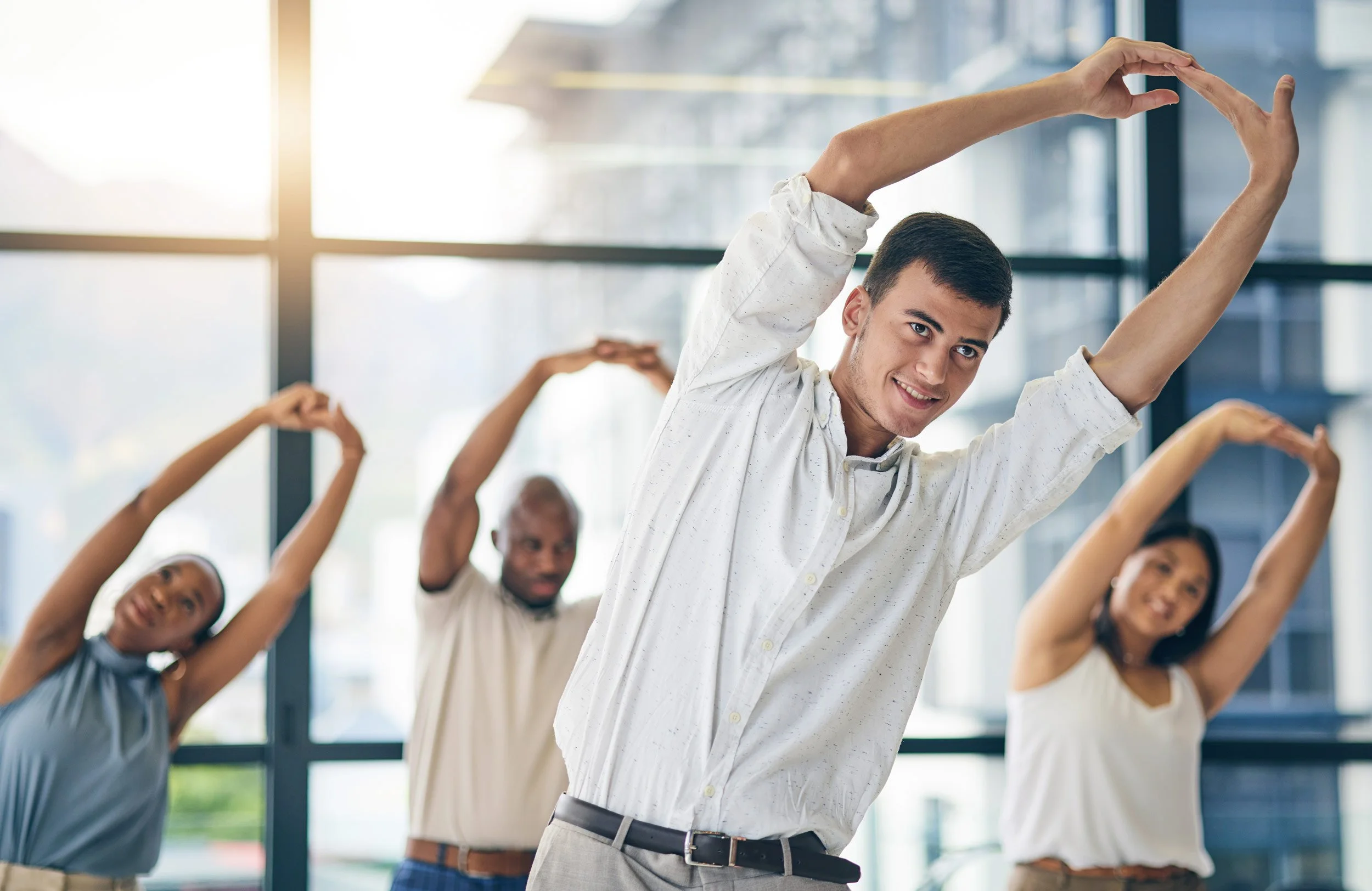 Gruppe von Menschen, die im Stehen in einem Raum mit großen Fenstern Yoga- oder Dehnübungen machen.