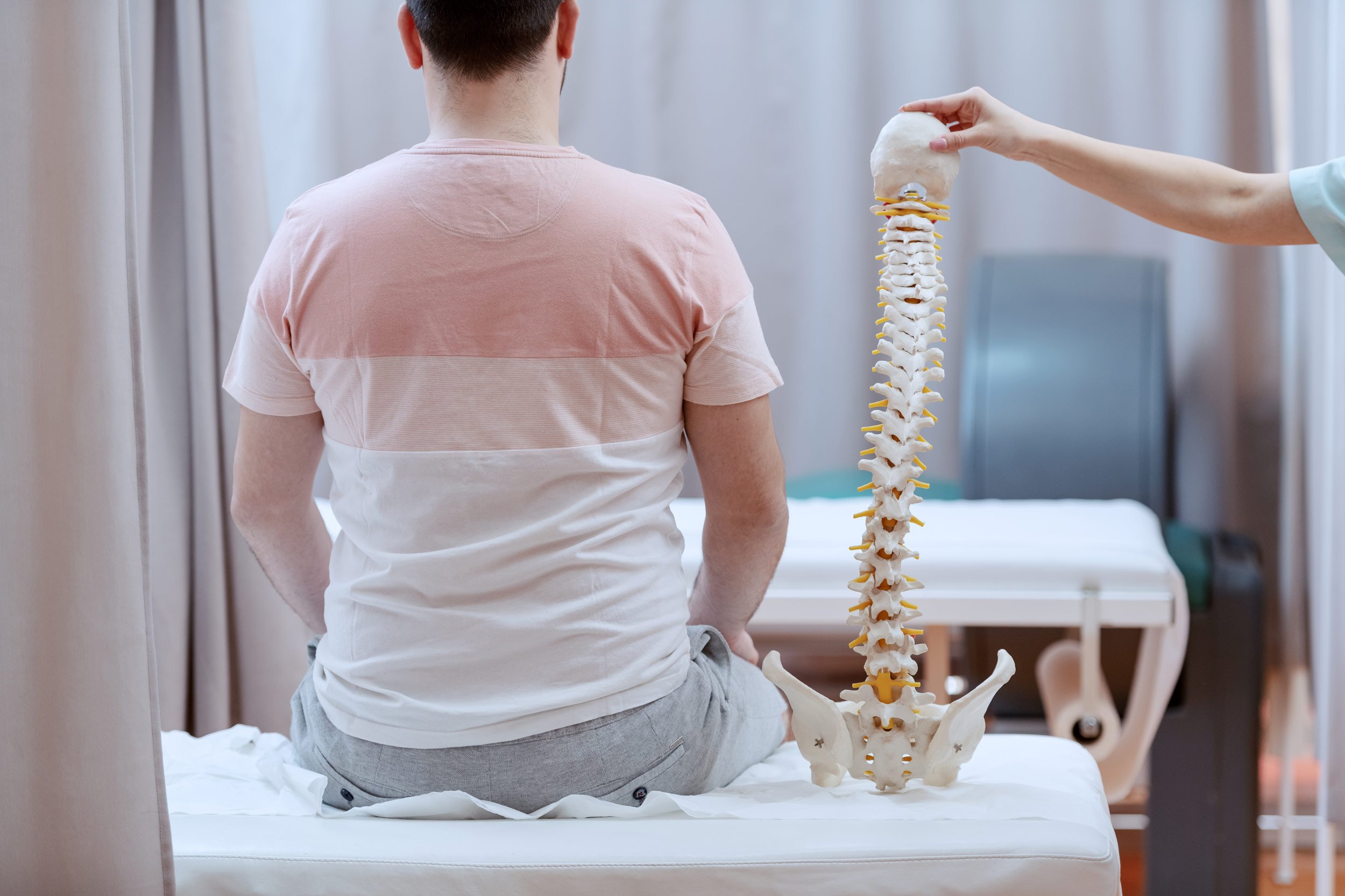 Ein Mensch mit kurz dunklem Haarschnitt sitzt auf einer Untersuchungsliege und blickt auf ein anatomisches Skelett der Wirbelsäule, das vor ihm steht. Im Hintergrund sind weiße Vorhänge.