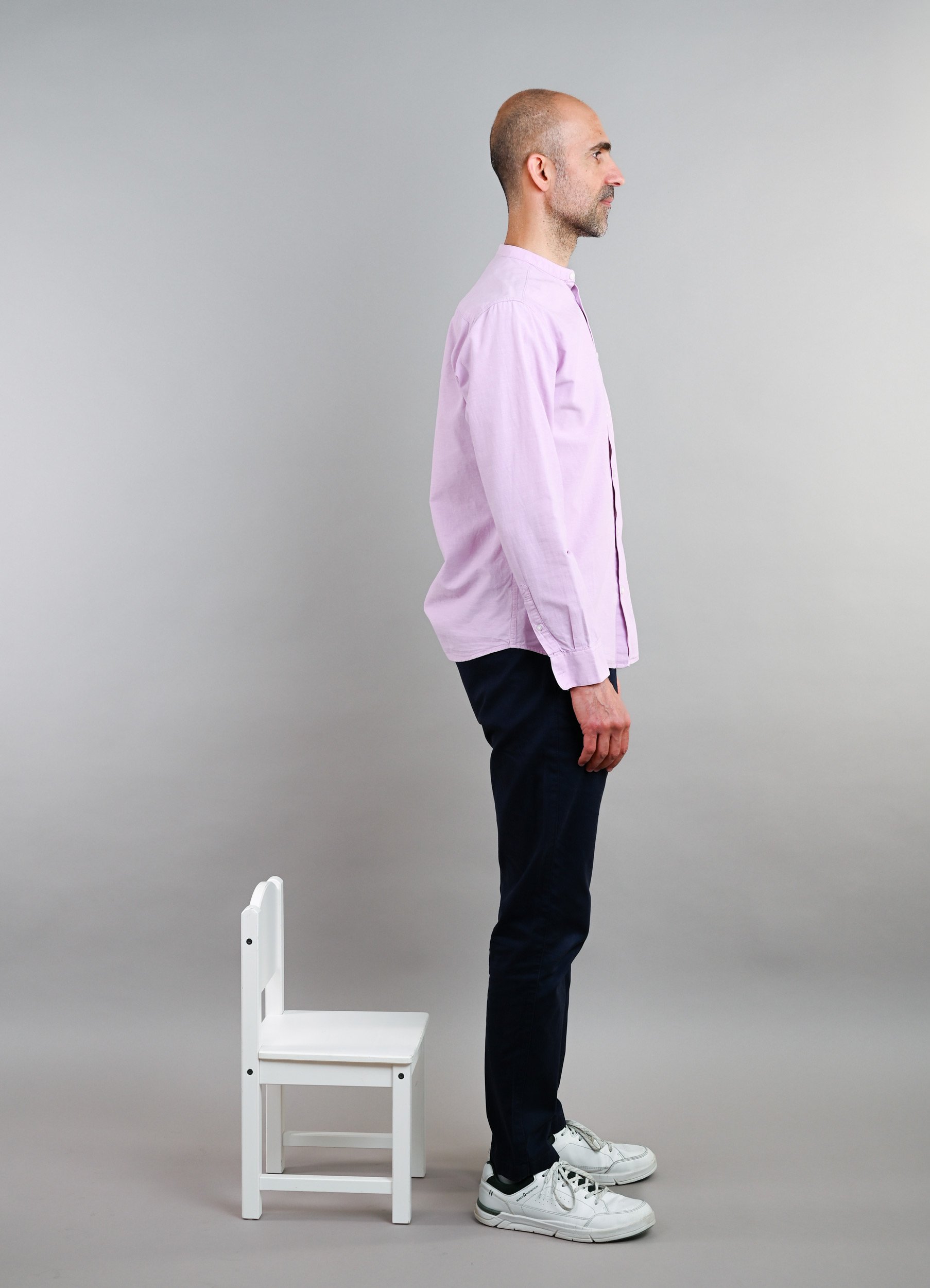 Ein Mann steht seitlich neben einem weißen Stuhl, der auf dem Boden steht. Der Mann trägt ein pinkfarbenes Hemd, dunkle Hosen und weiße Turnschuhe. Der Hintergrund ist grau.
