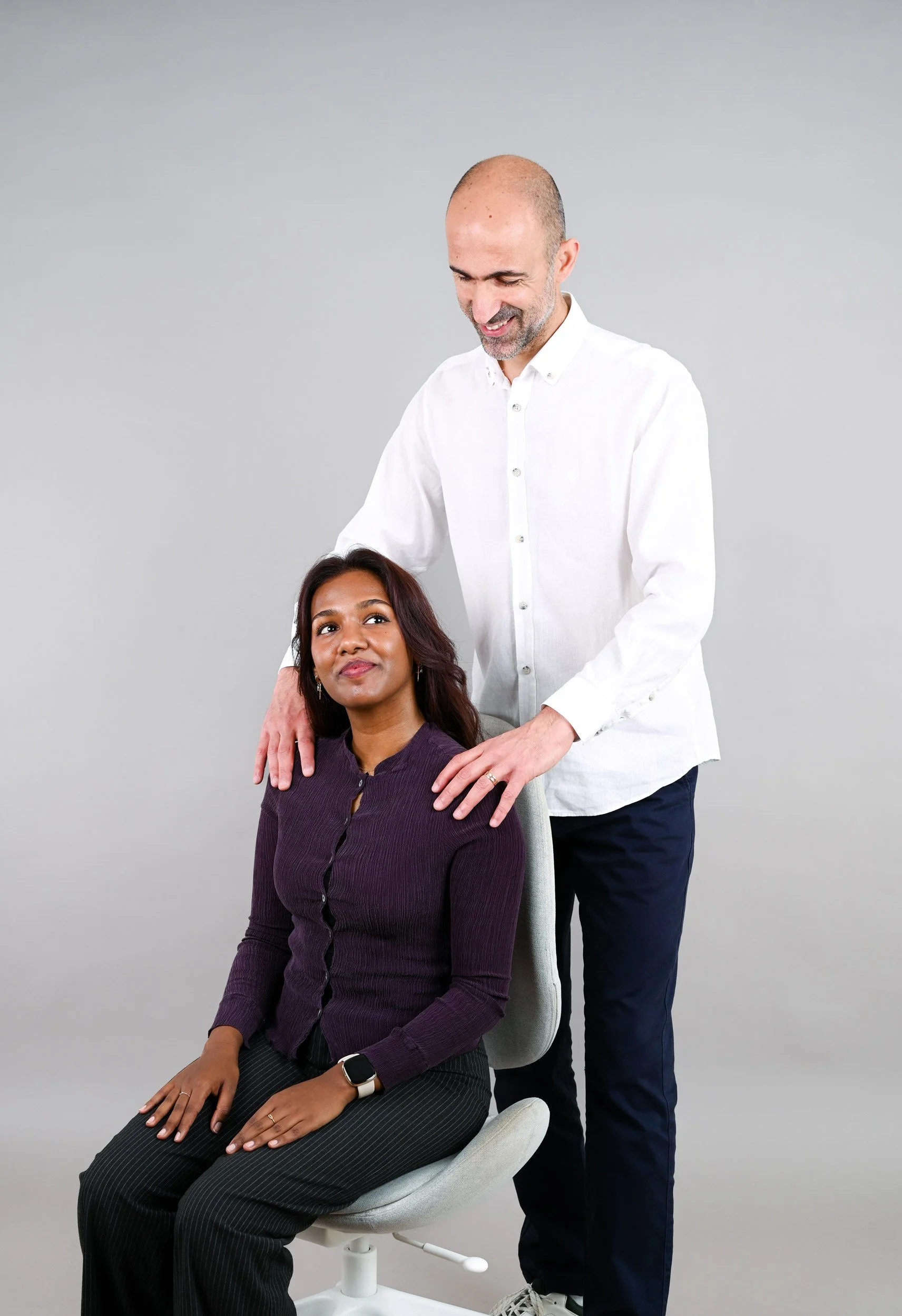 Ein Mann arbeitet an einer Frau, die auf einem Stuhl sitzt, im medizinischen oder therapeutischen Setting, mit neutralem grauem Hintergrund.