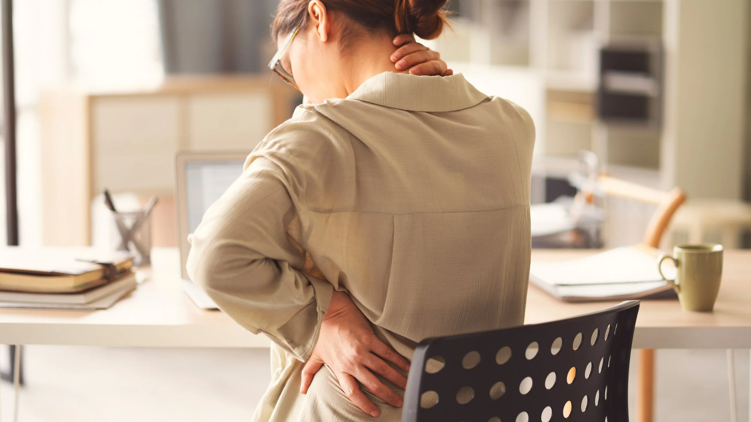 Frau hält sich am Nacken und Rücken, steht an einem Schreibtisch in einem Büro.
