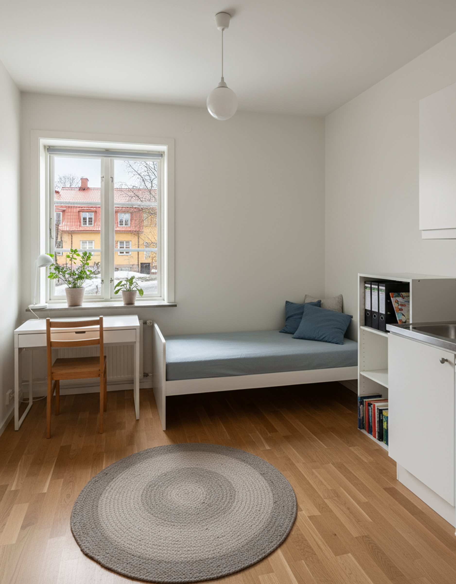 Ett minimalistiskt rum med ett sängstativ, en säng med blå lakan och kuddar, en vit byrå med balkingsfiler, ett fönster med utsikt över en gata utomhus, ett papperskuvert på golvet, och en liten arbetsstation vid fönstret med två växter på fönsterbrädan.