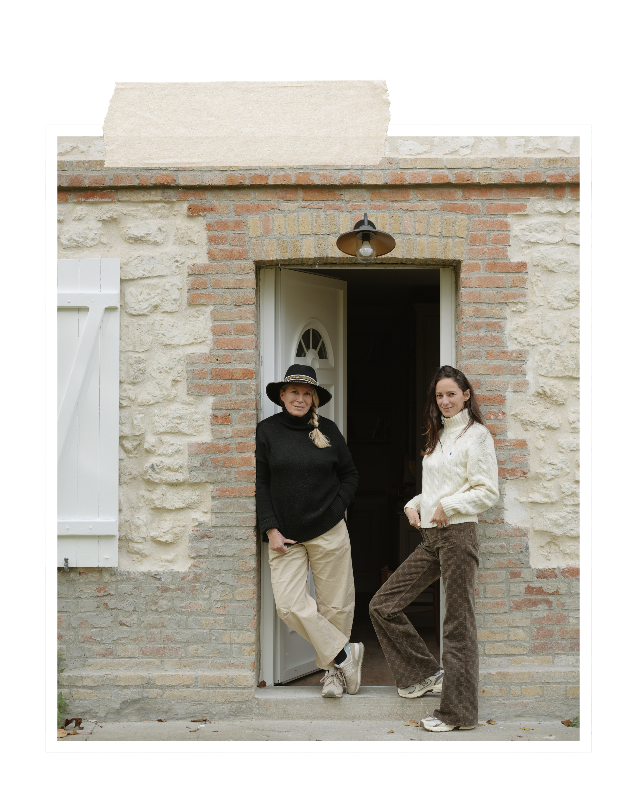 Two women standing at the doorway of a brick house with stone and brick exterior, open door, and a light fixture above the door, one in black sweater and hat, the other in white sweater and patterned pants.