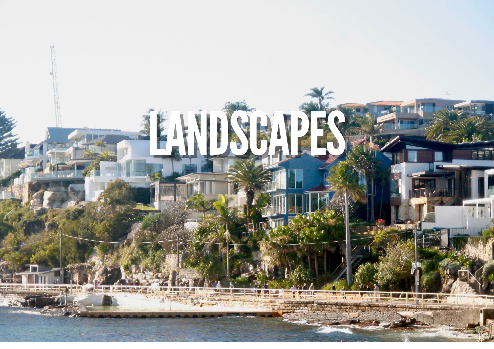 A coastal scene with modern houses on a hillside surrounded by palm trees, overlooking the water with a promenade and people walking along the shore.
