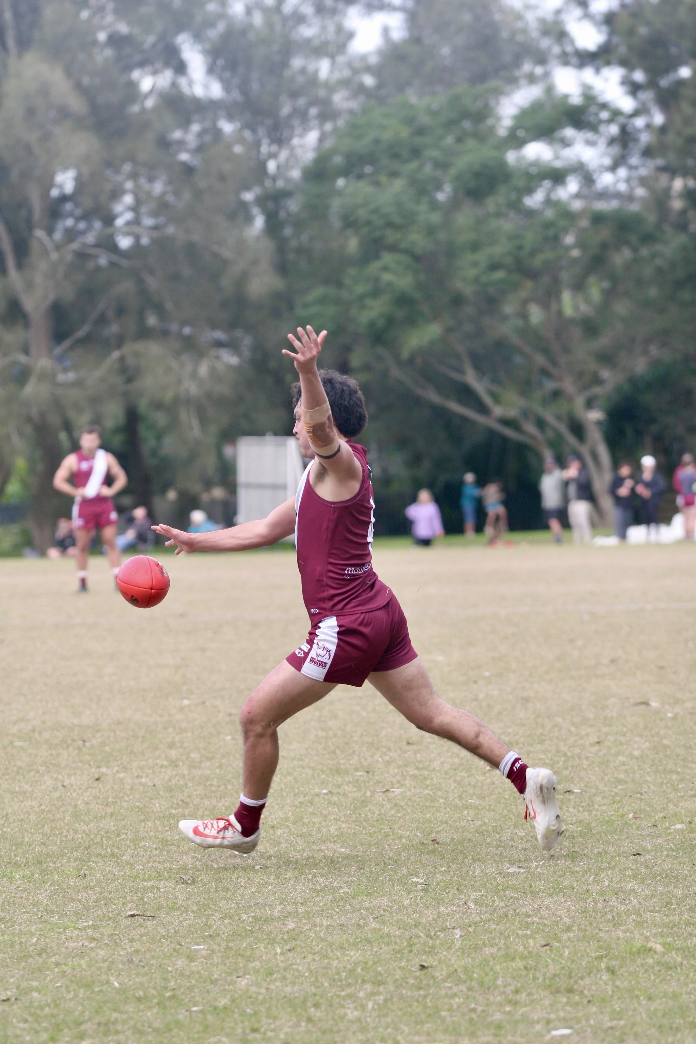 A person in maroon sports uniform kicking a red football on a grassy field during daytime, with people and trees in the background.