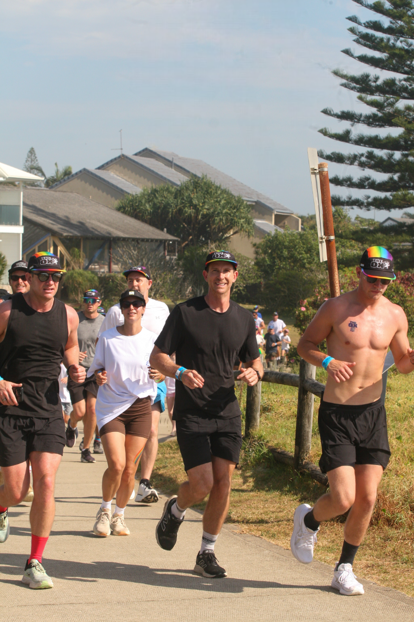 Group of runners participating in an outdoor race on a sunny day, some wearing black shirts and shorts, others in white or without shirts, with houses and trees in the background.