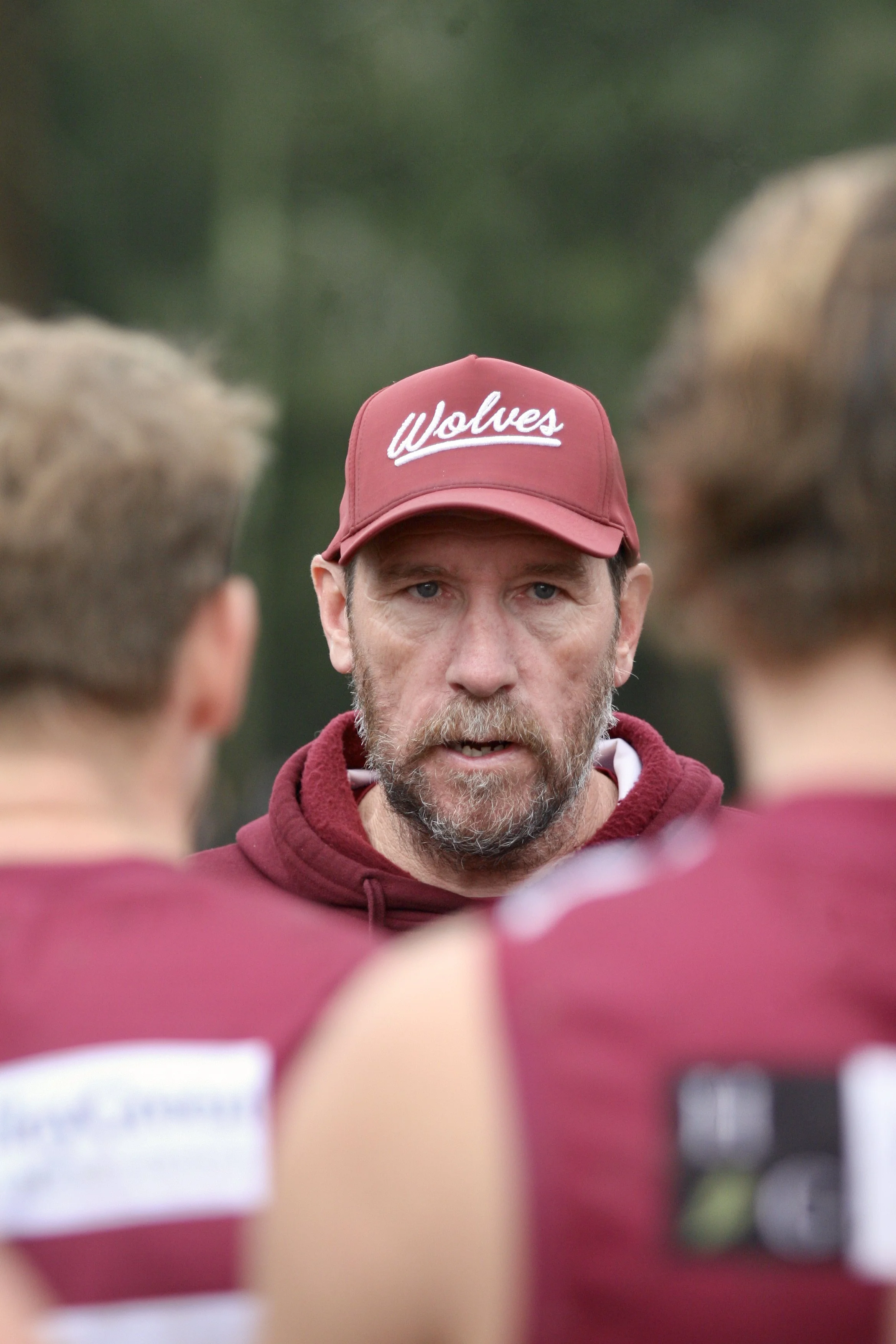 A coach with a beard and red cap labeled 'Wolves' speaking to football players in maroon uniforms during a game.