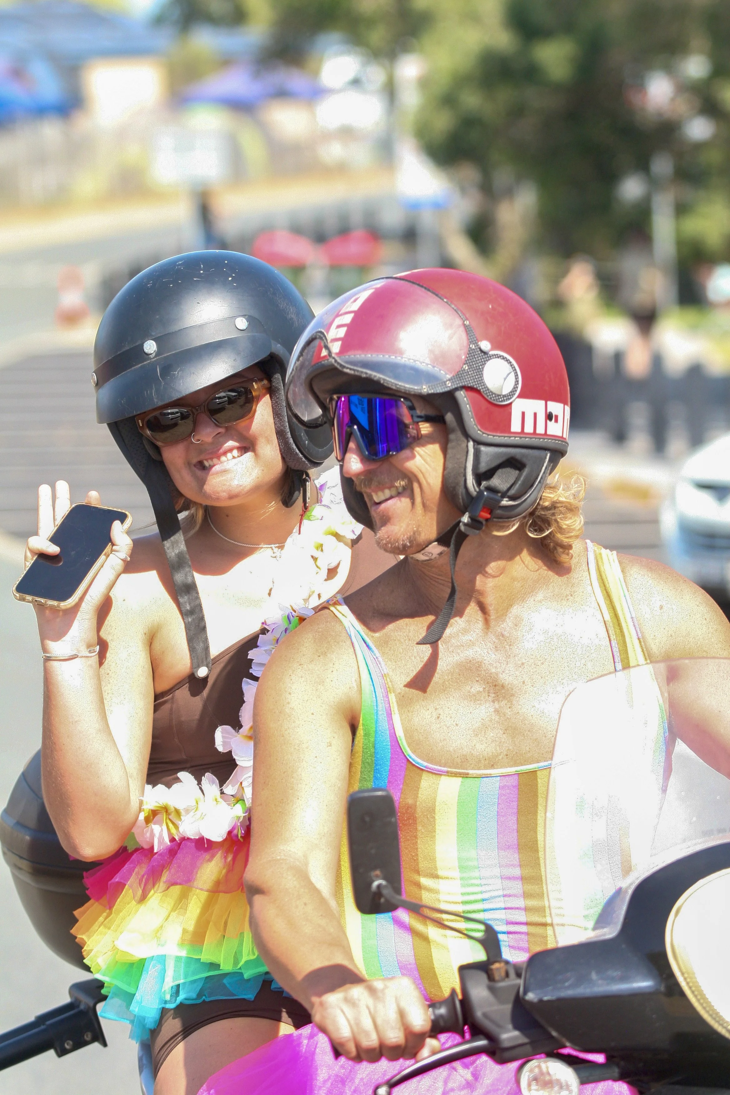 Two people, a man and a woman, riding a motorcycle or scooter outdoors on a sunny day wearing helmets, sunglasses, and colorful clothing. They are smiling and appear to be having fun.