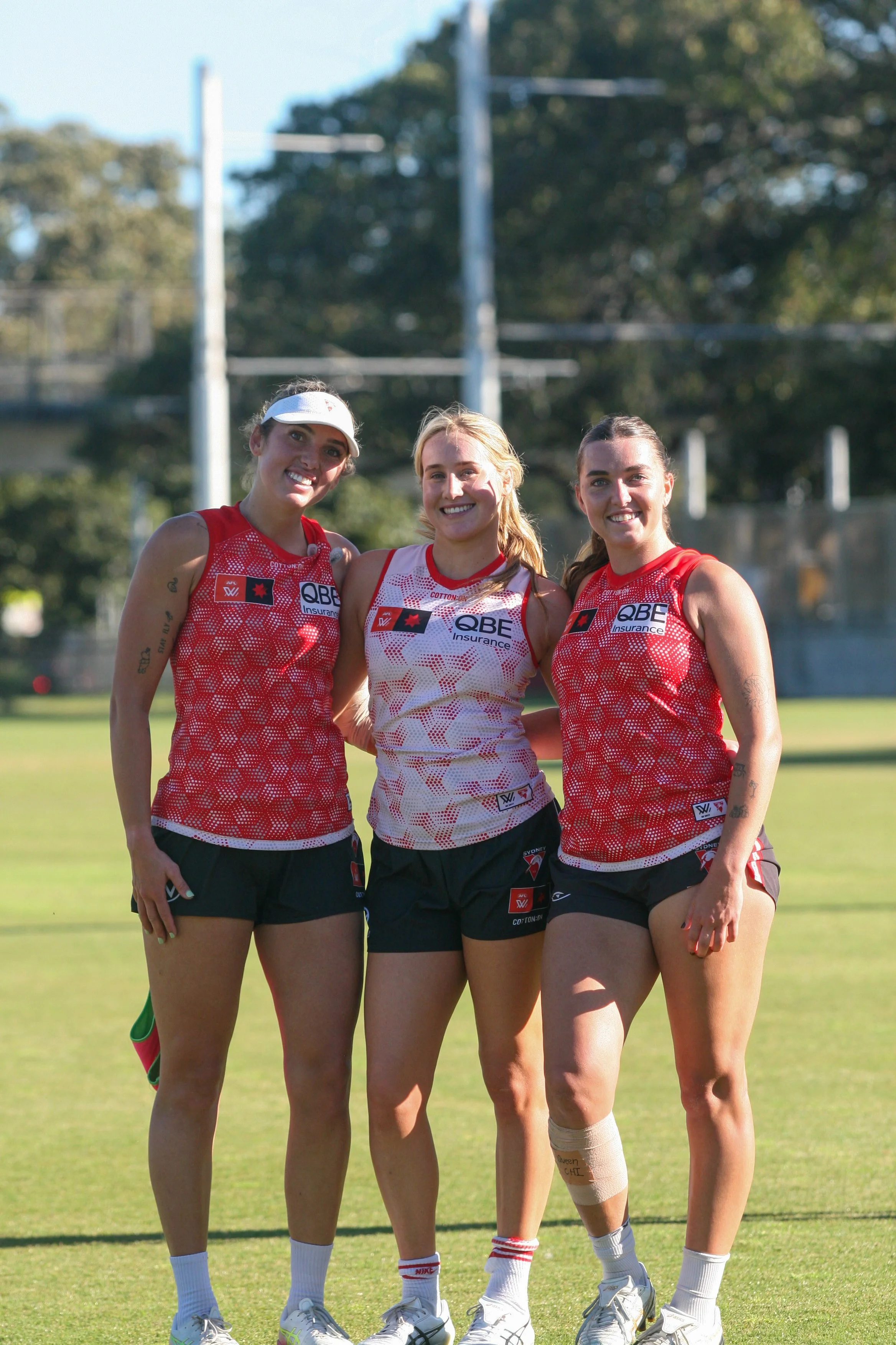 Three female Australian Rules football players in red and white jerseys and black shorts standing on a grass field, smiling at the camera.