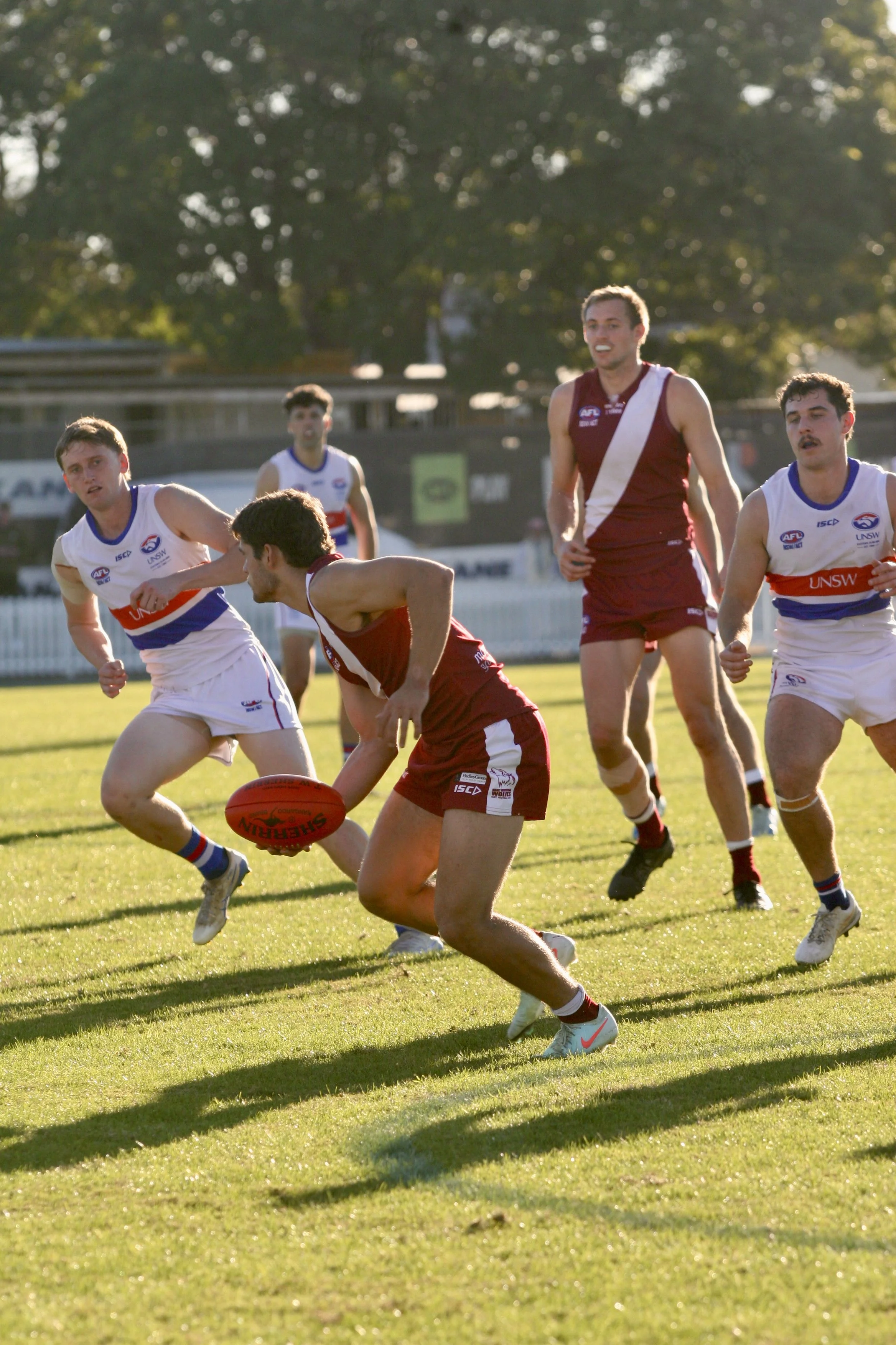 Australian rules football game with players in maroon and white uniforms competing on a grassy field during daytime.
