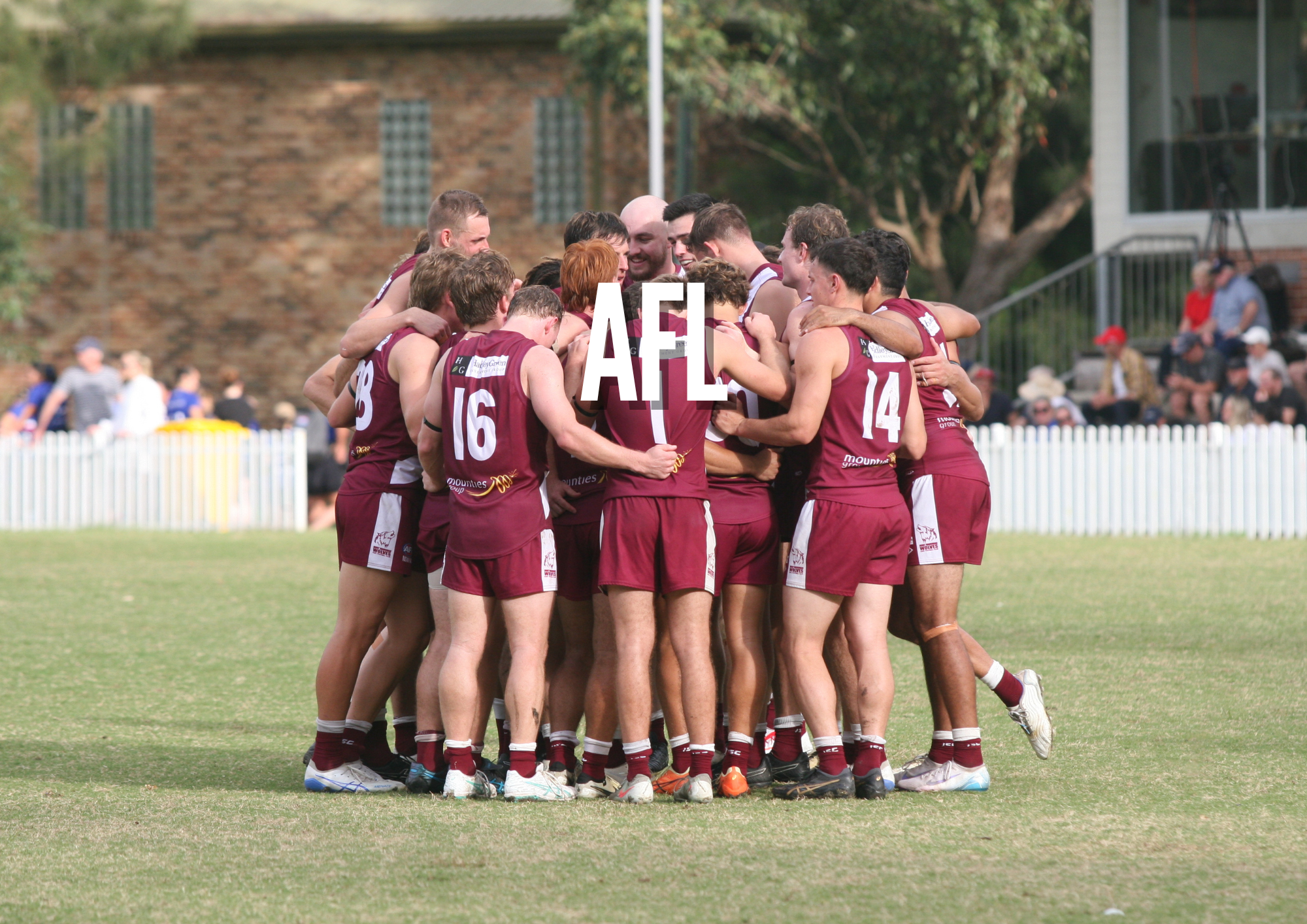 Australian rules football team huddled together on the field celebrating a team moment with spectators in the background.