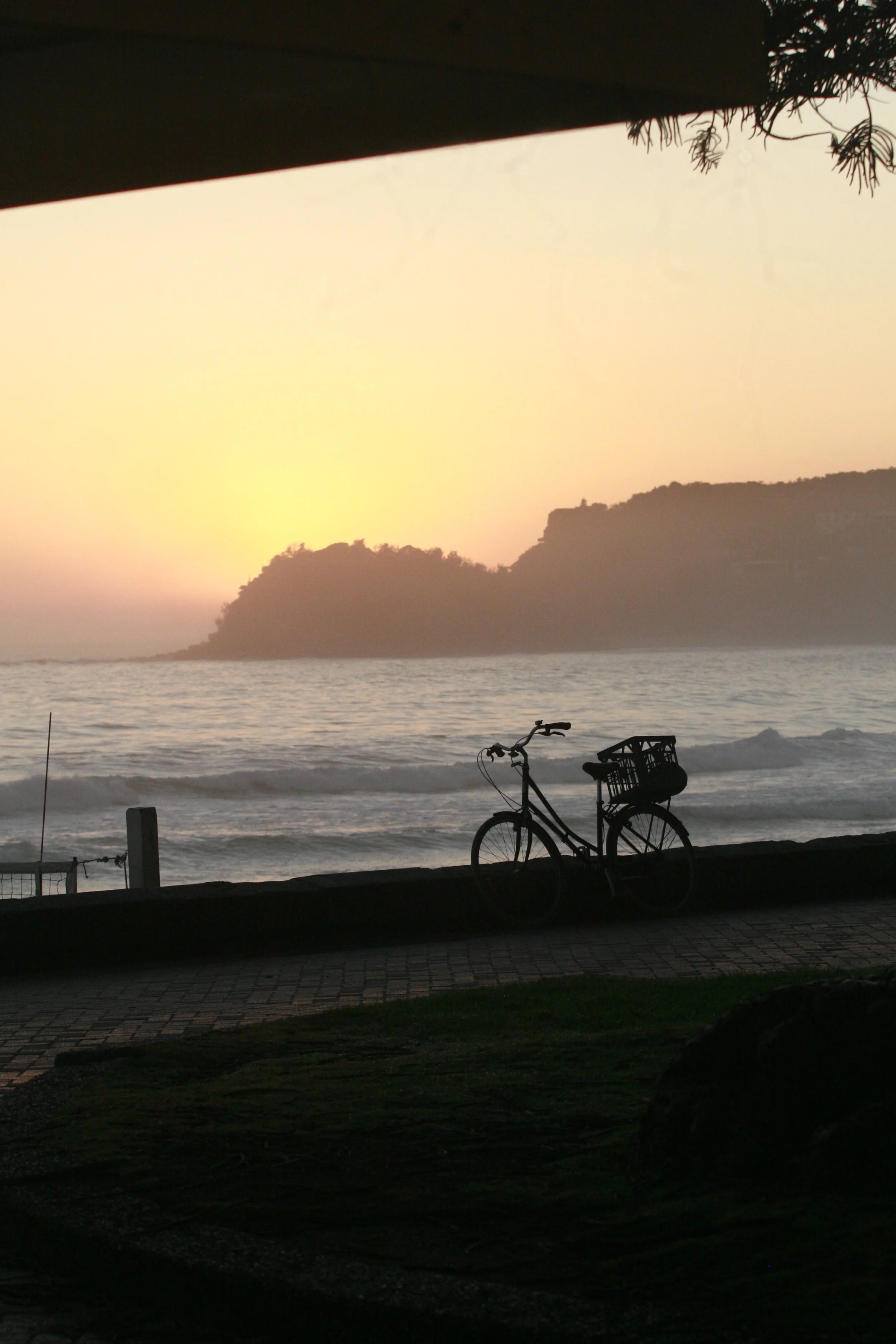Silhouette of a bicycle with a basket on a beach promenade at sunset, with ocean waves and distant hills in the background.