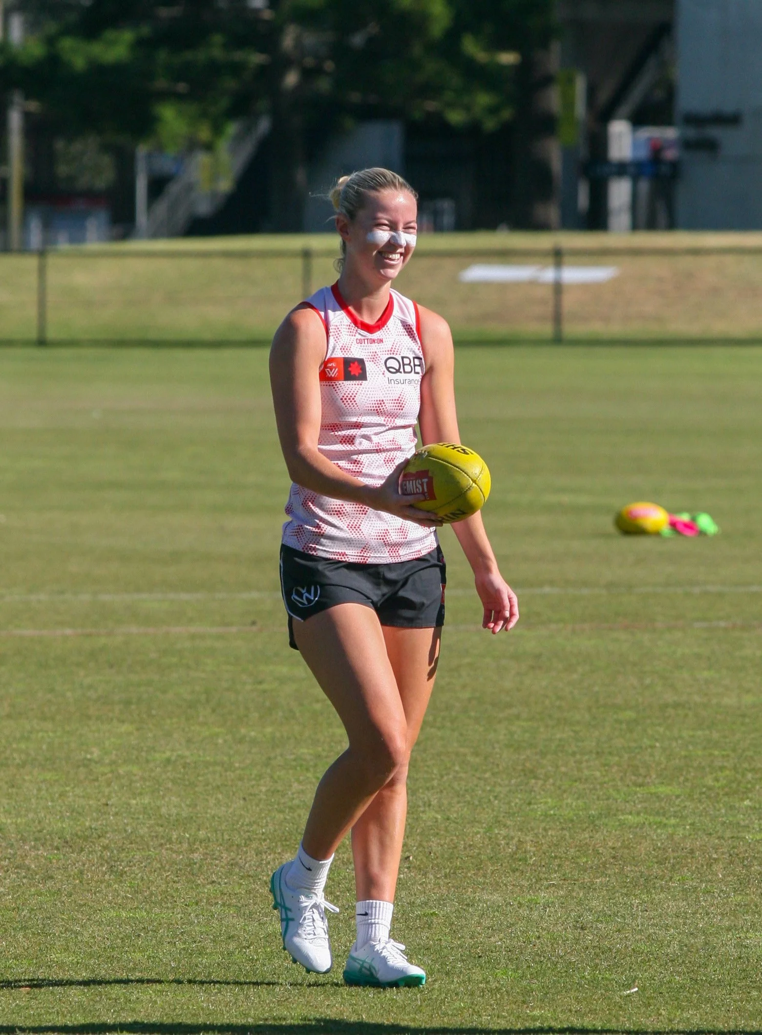 A woman wearing athletic clothing and white sneakers is holding a yellow football on a grass field. She is smiling and appears to be playing or practicing in a sports setting.