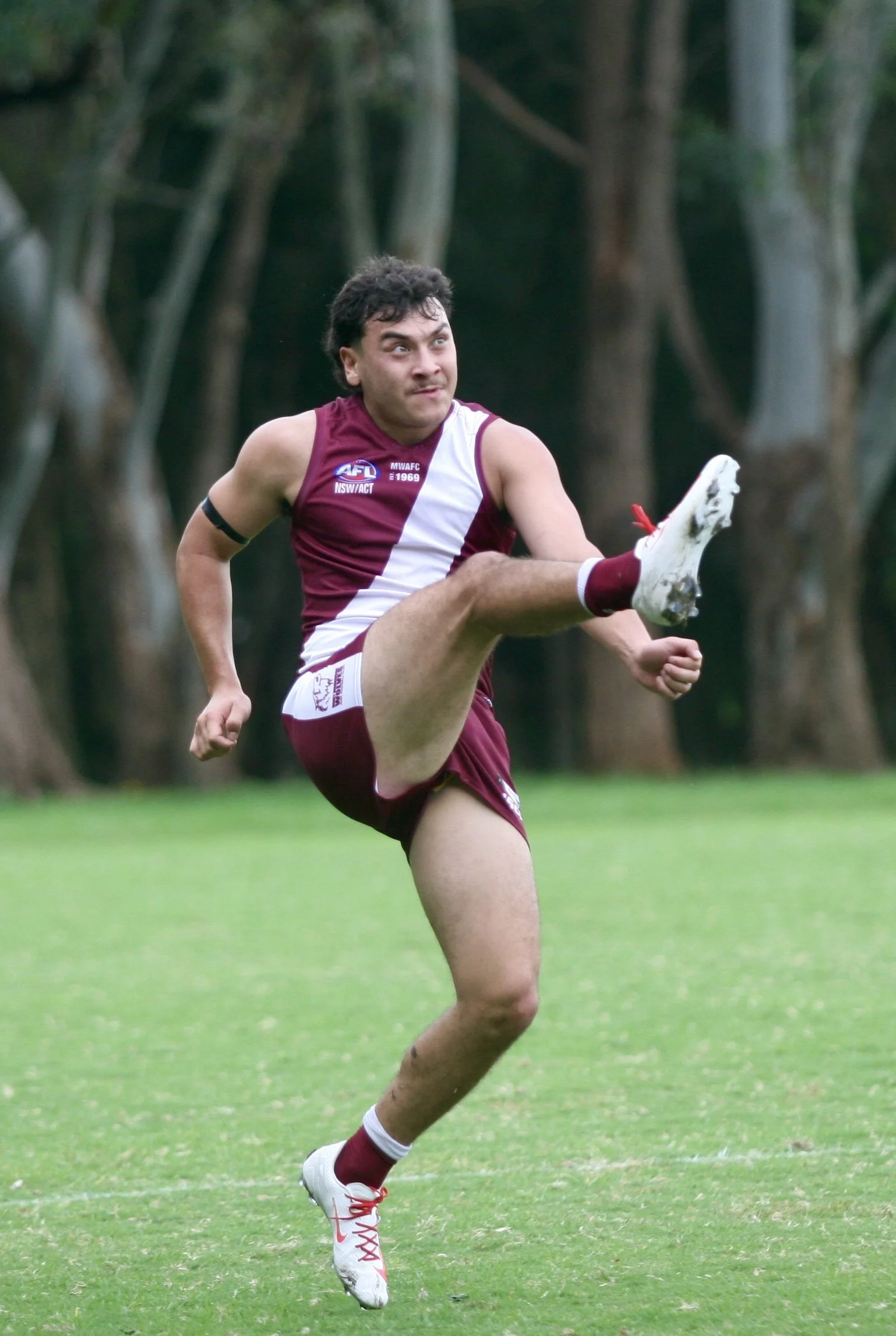 A man in a maroon and white sports uniform kicking a football on a grassy field with trees in the background.