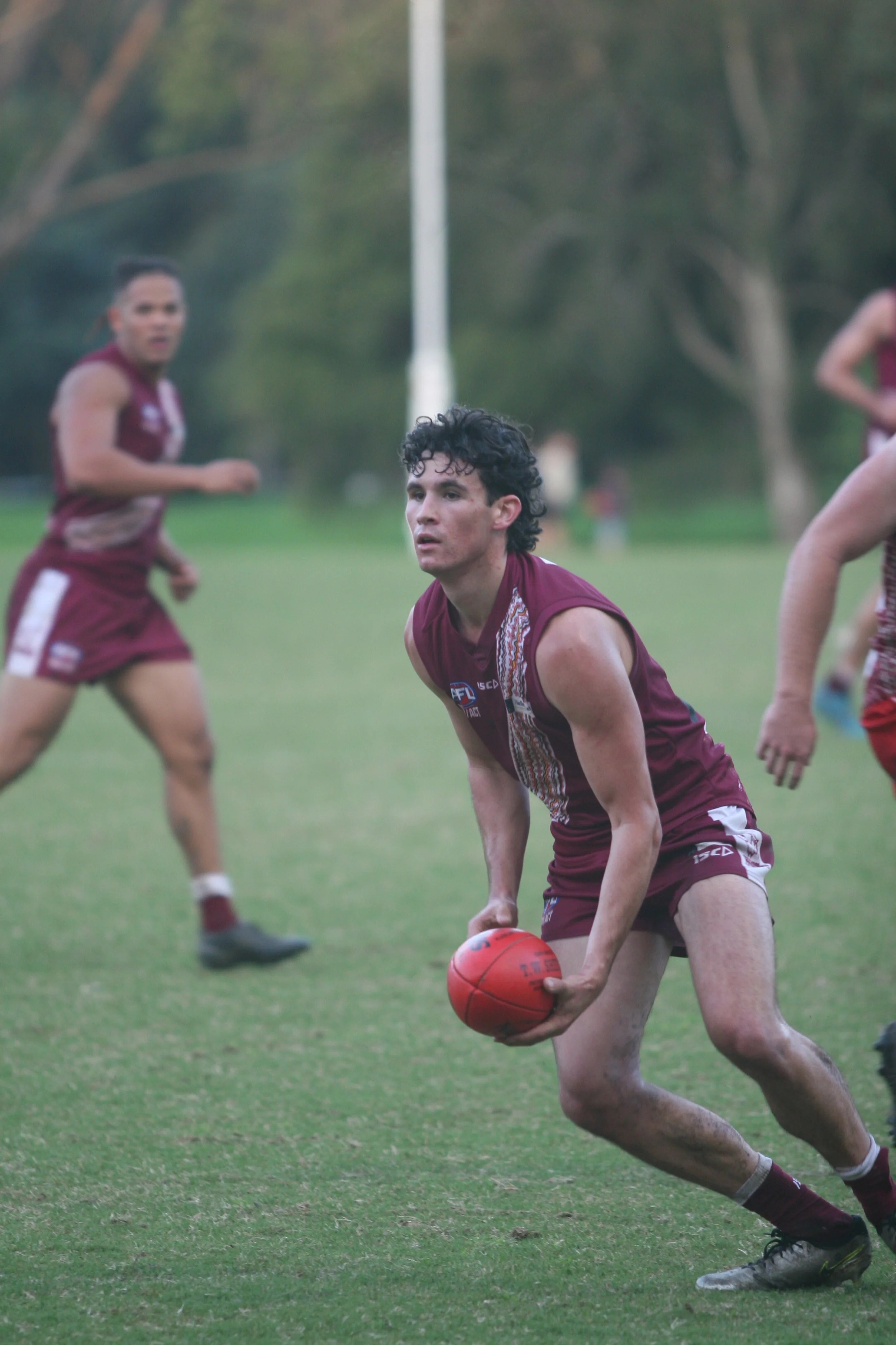 A male Australian rules football player in a maroon uniform holding a red football on a grassy field, with other players in the background.