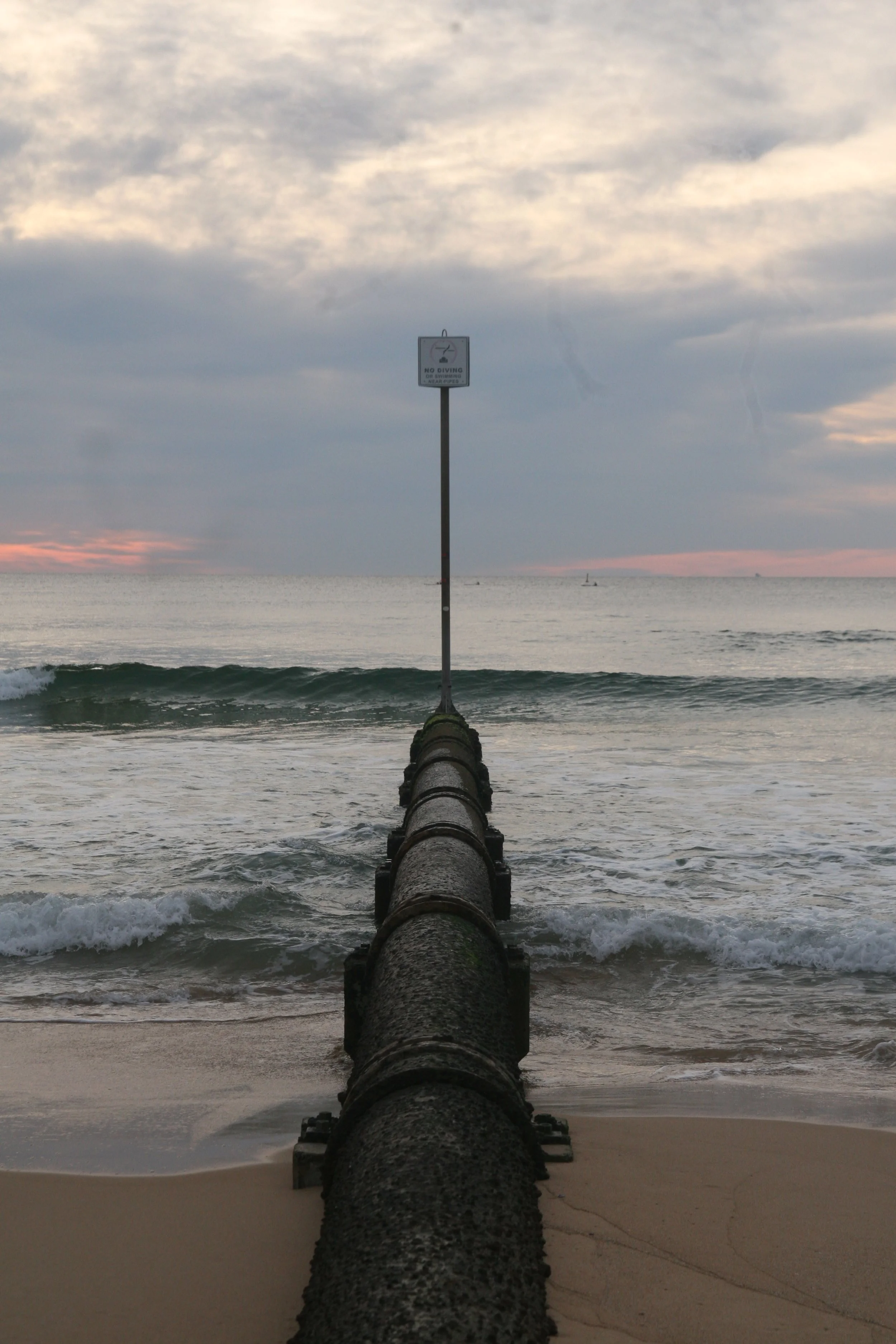 A large pipe extending into the ocean with a 'No Diving' sign at the end of a pole on top of the pipe, at the beach during sunset.