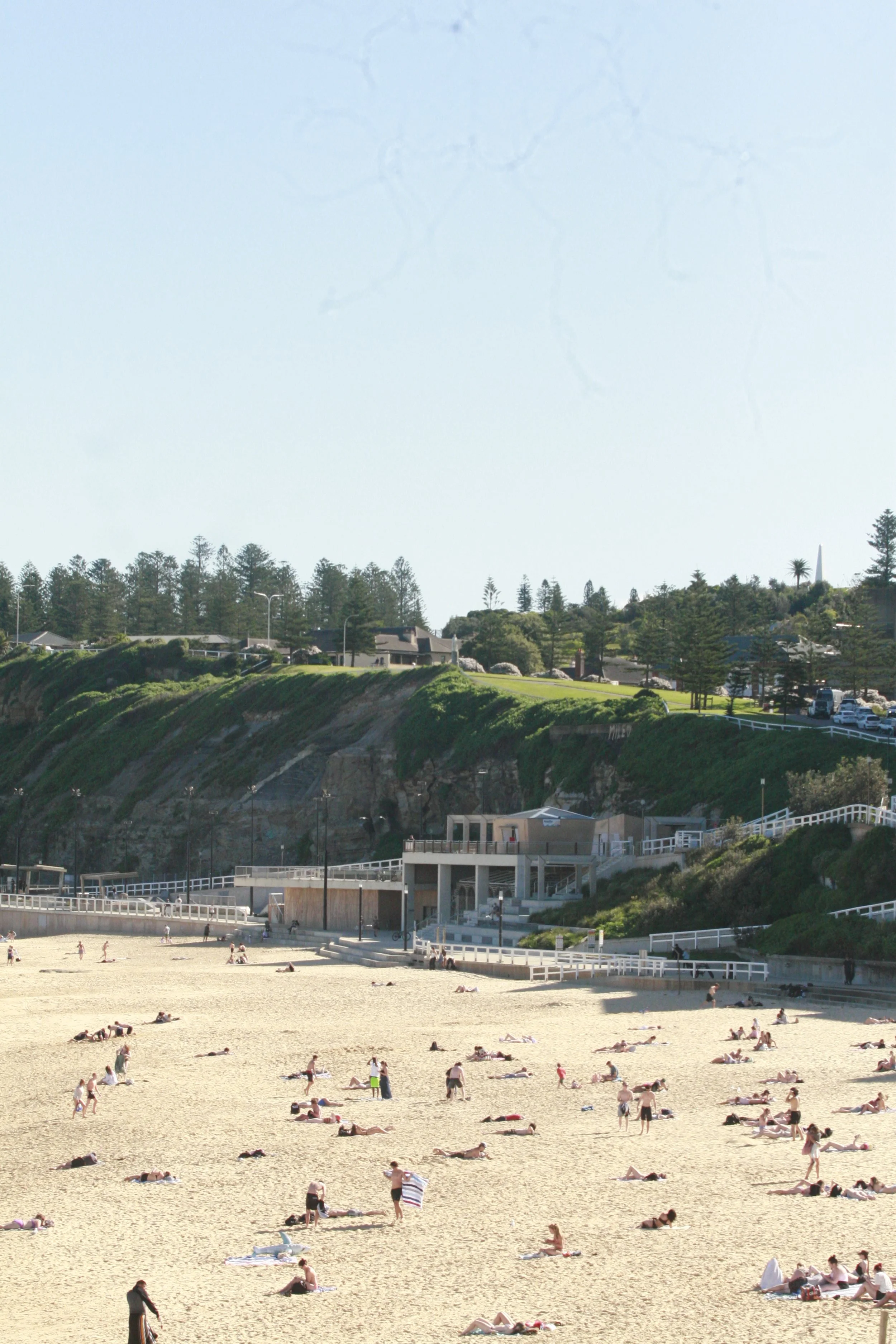 People sunbathing and relaxing on a sandy beach with a rocky cliff and green hillside in the background.