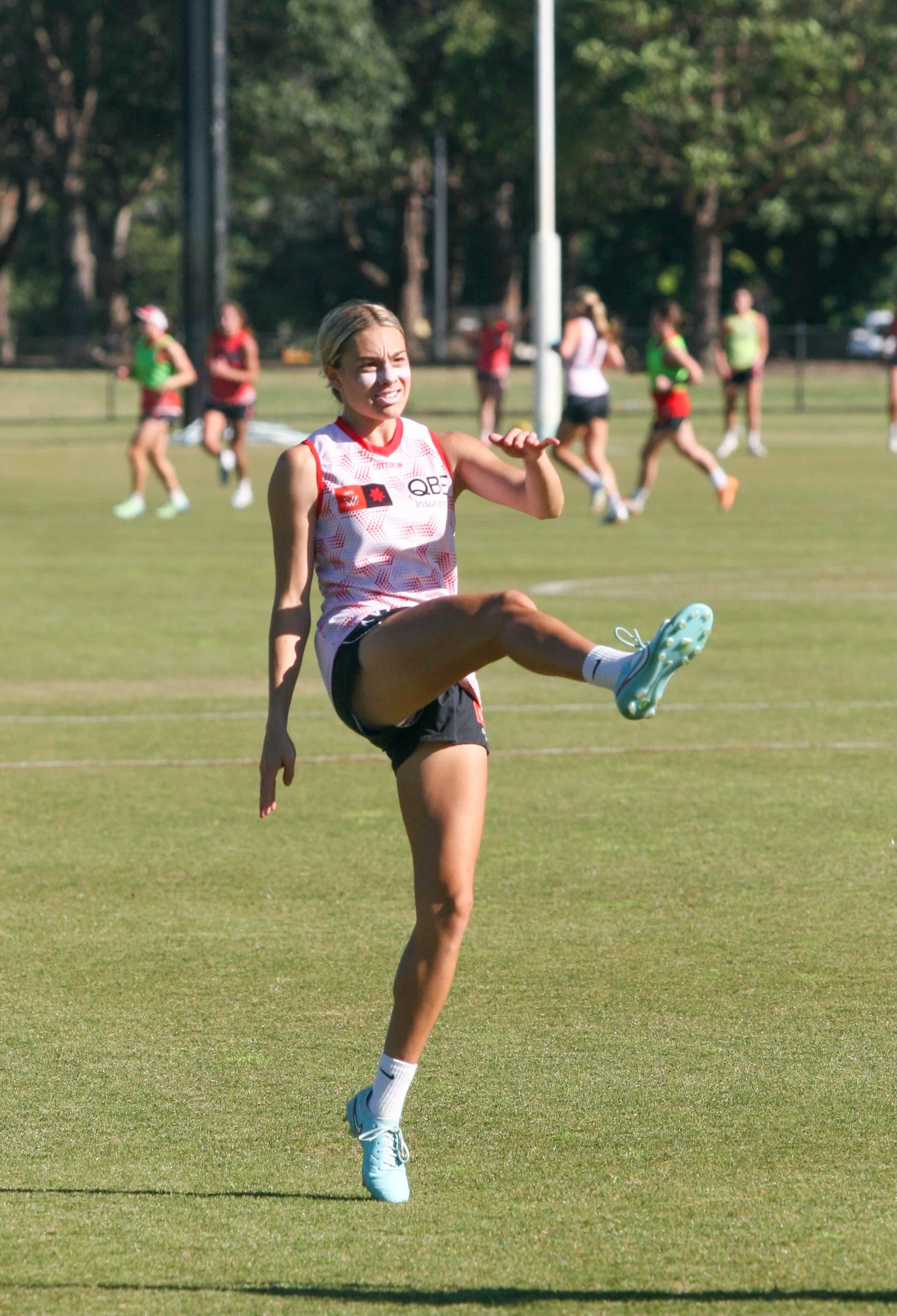 Female athlete on a sports field practicing kicking, with other players running in the background.