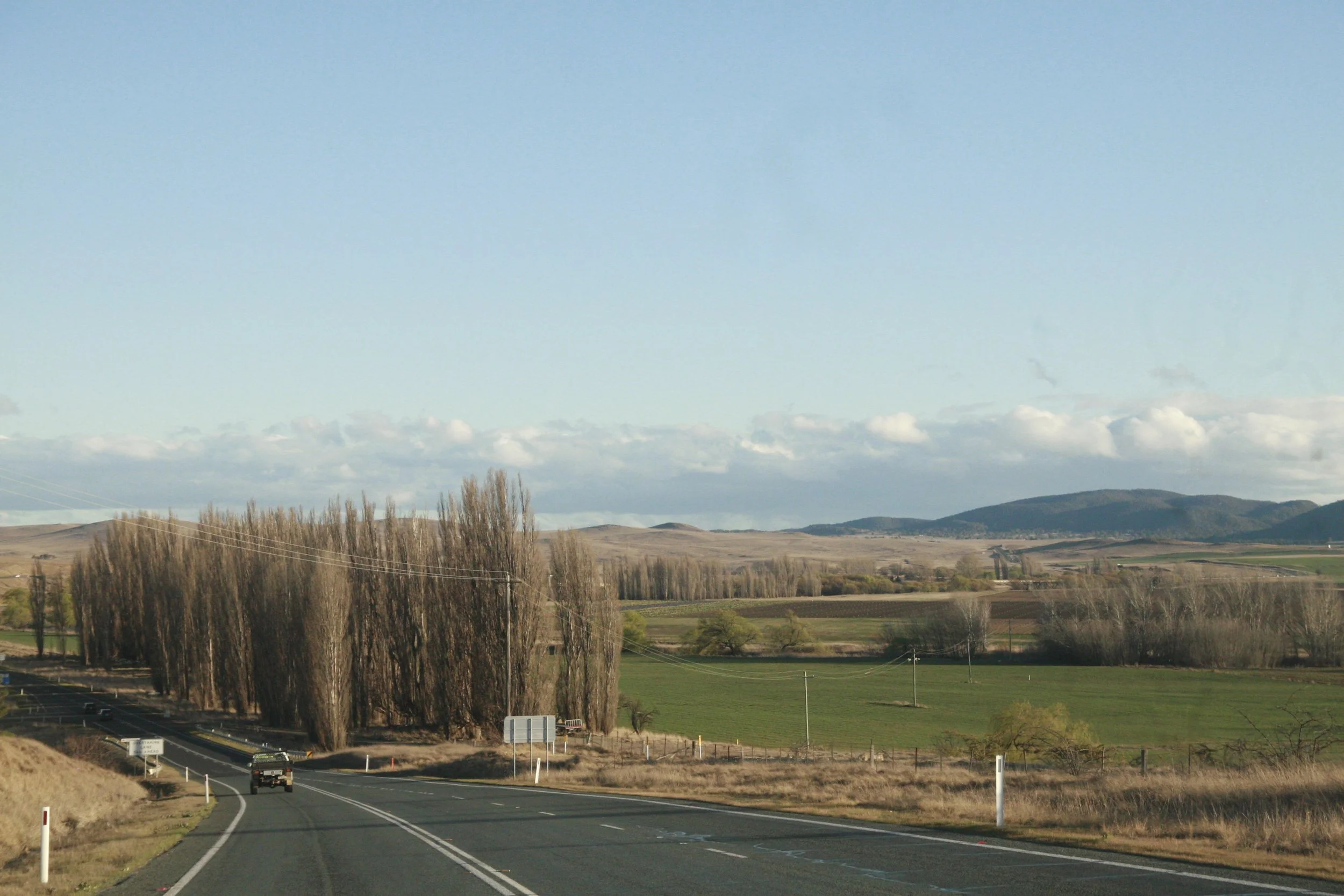 A two-lane rural highway with a truck driving in the distance, surrounded by fields, leafless trees, and rolling hills under a partly cloudy sky.