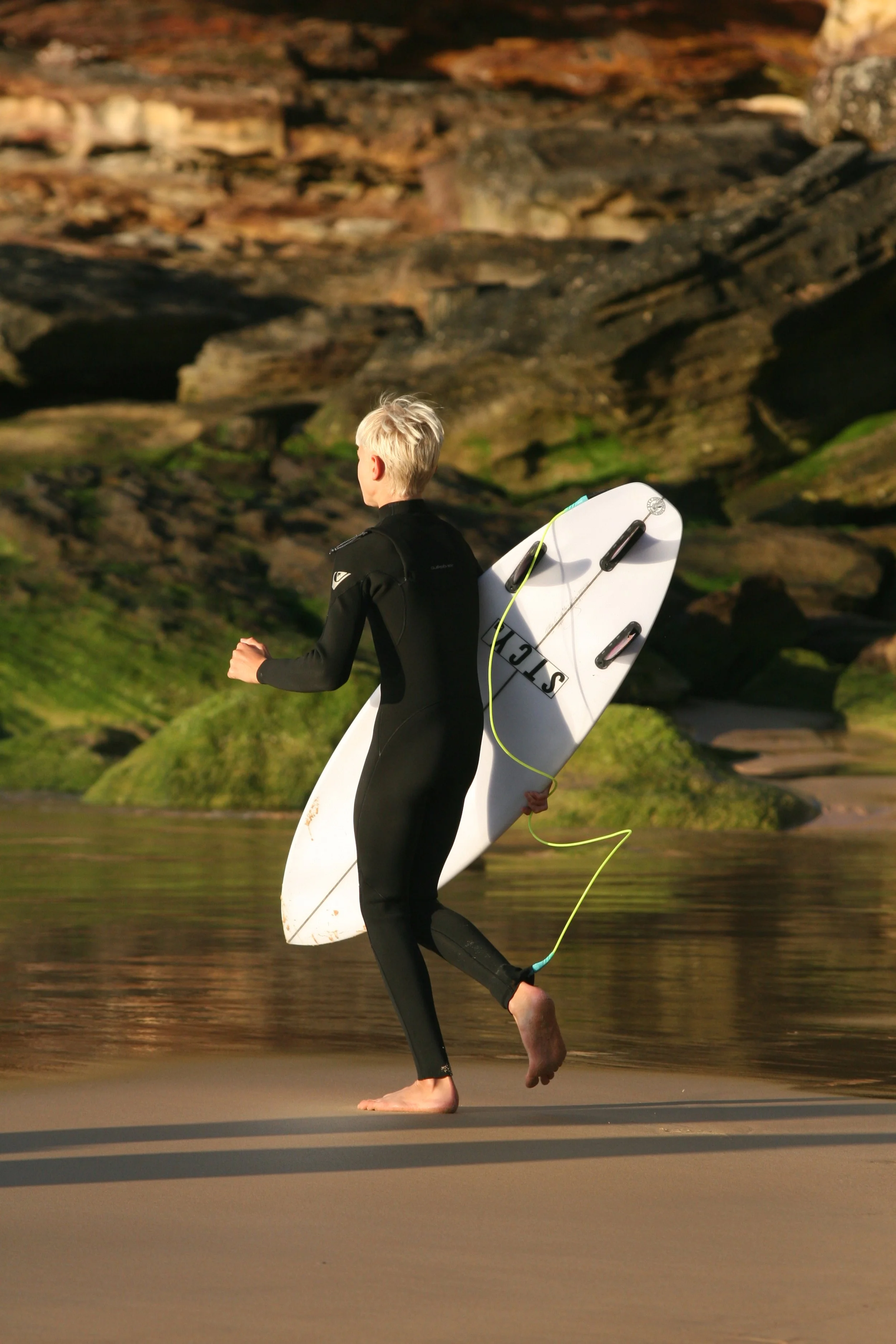 Person in a wetsuit carrying a surfboard walking on the beach near the water with rocky cliffs in the background.