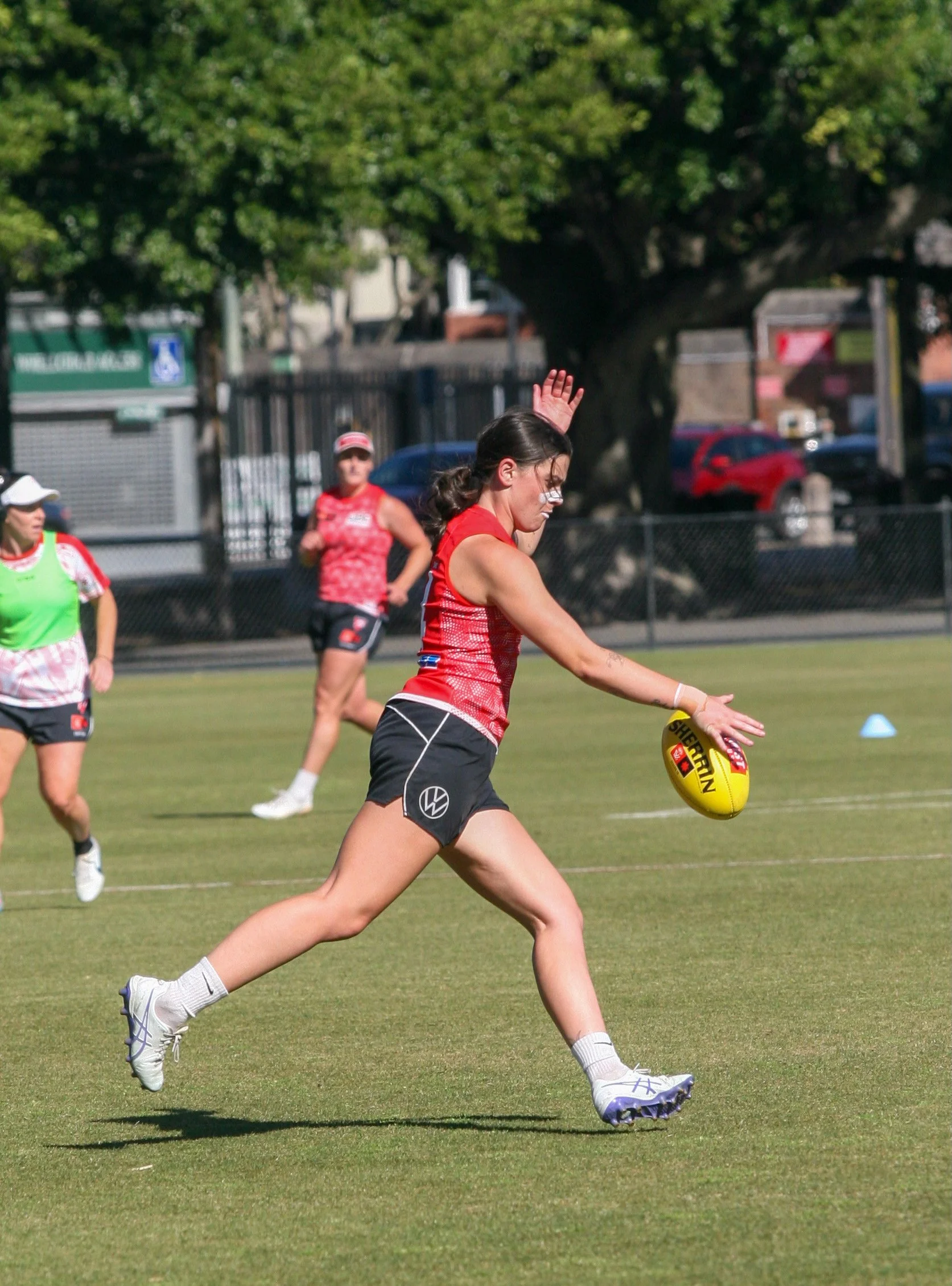 A female athlete dressed in a red and black sports uniform is running on a grassy field, about to kick a yellow Australian rules football. Other players and trees are visible in the background.