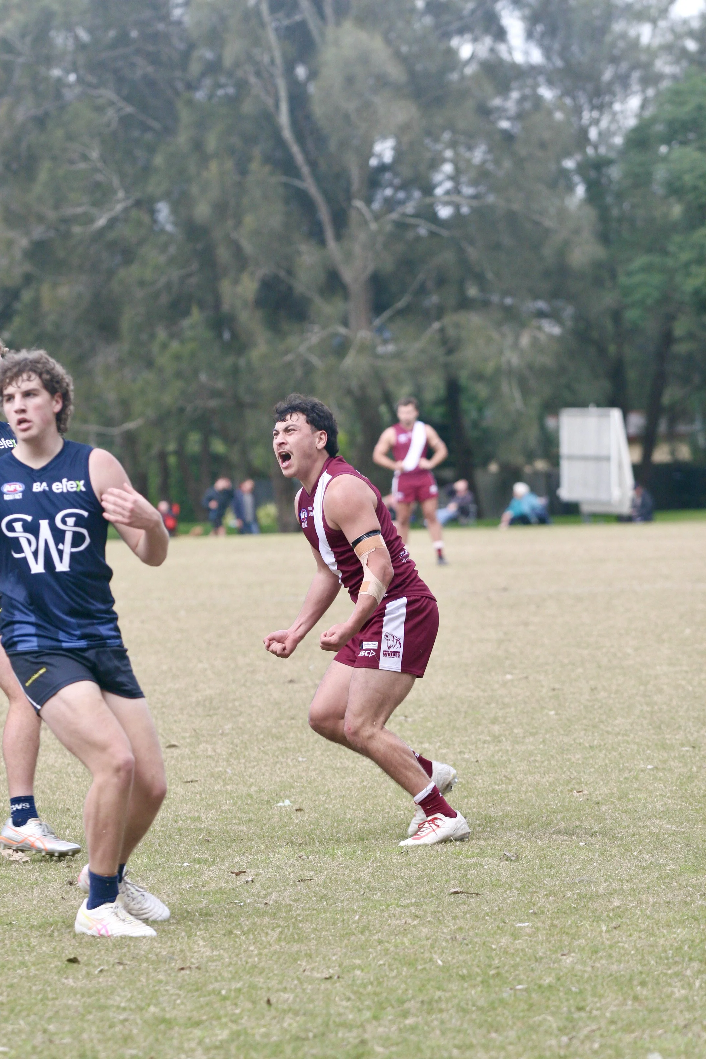 A rugby player in maroon uniform passionately celebrating on the field with clenched fists, while a player in a blue jersey looks on. Other players and spectators are visible in the background with trees surrounding the field.