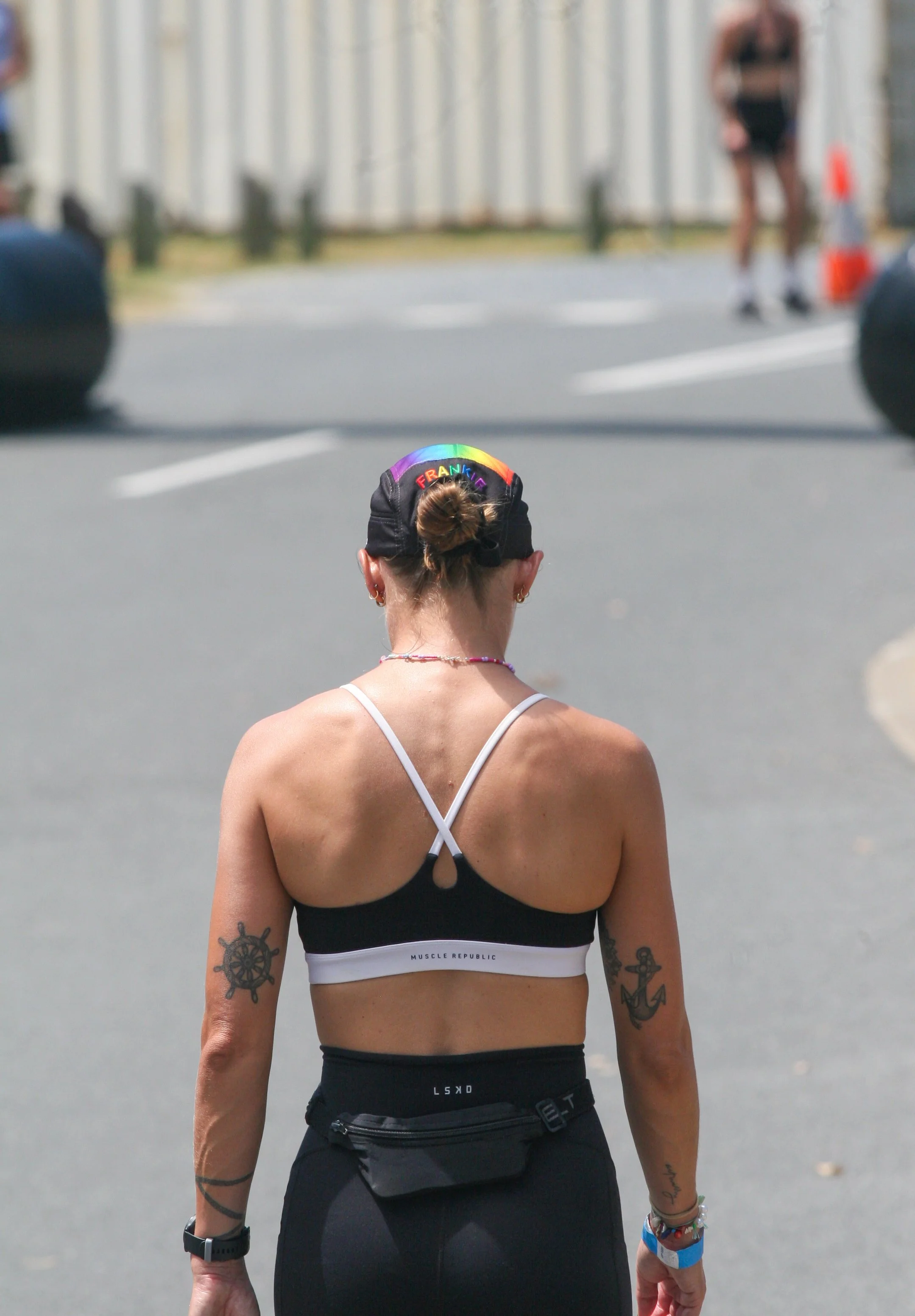 Woman with tattoos, wearing a black sports bra and black leggings, walking outdoors during the day.