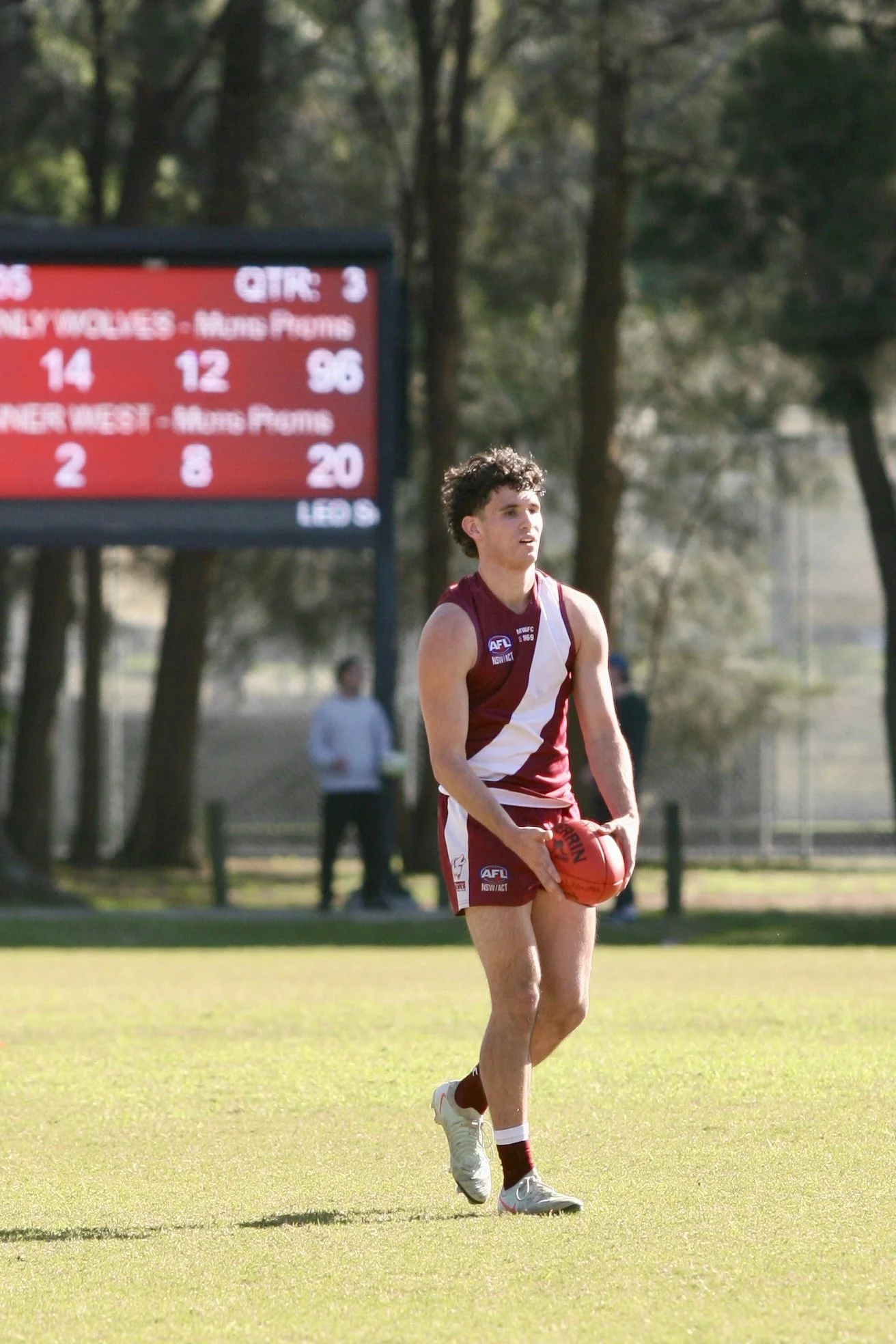 A young male Australian Football League player standing on a football field, holding a red football, wearing a maroon and white uniform, with a scoreboard and trees in the background.