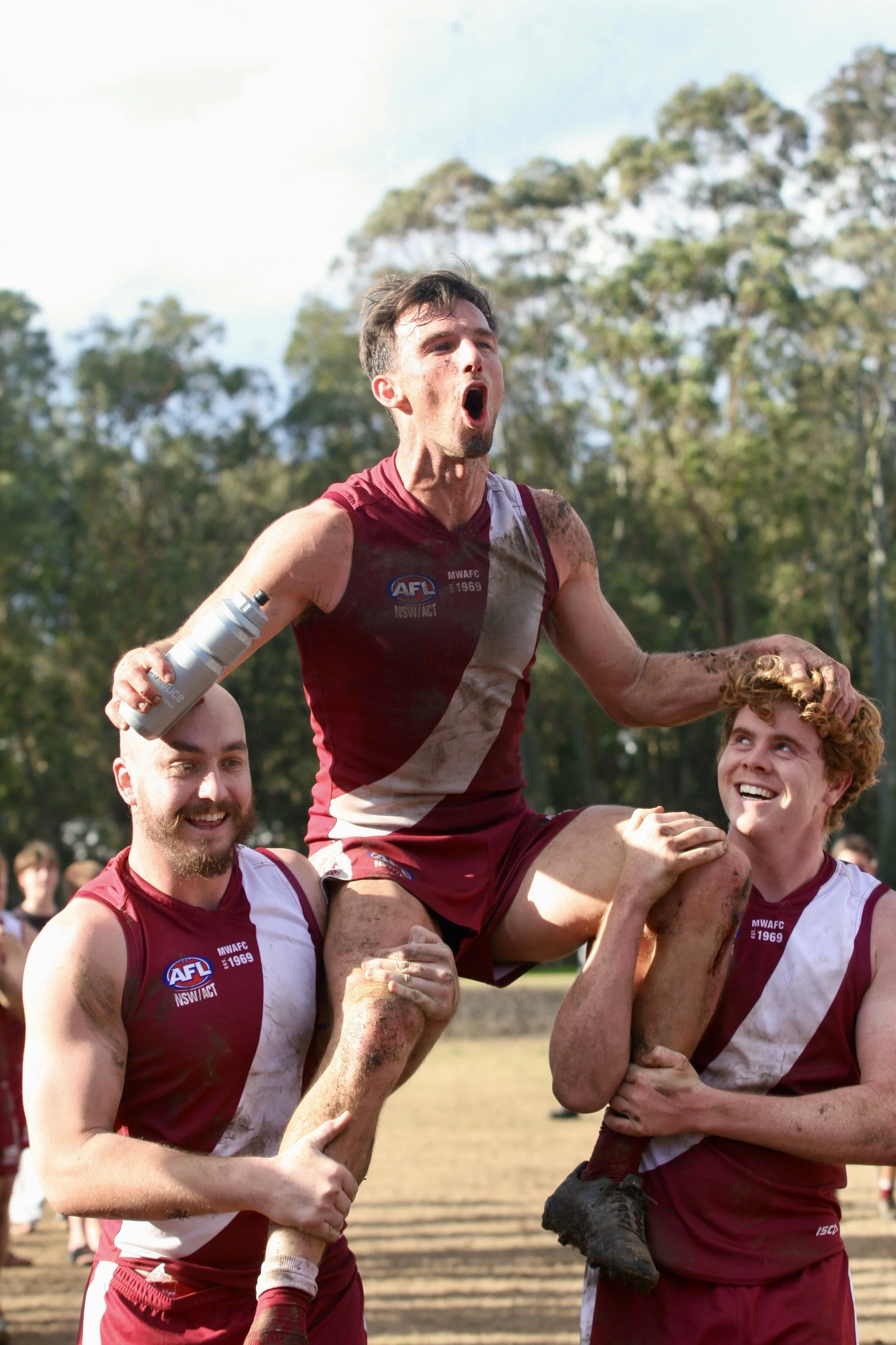 Australian football players celebrating after a match, lifting one player on top of two others. The players are wearing maroon and white uniforms covered in dirt, with a wooded area in the background.