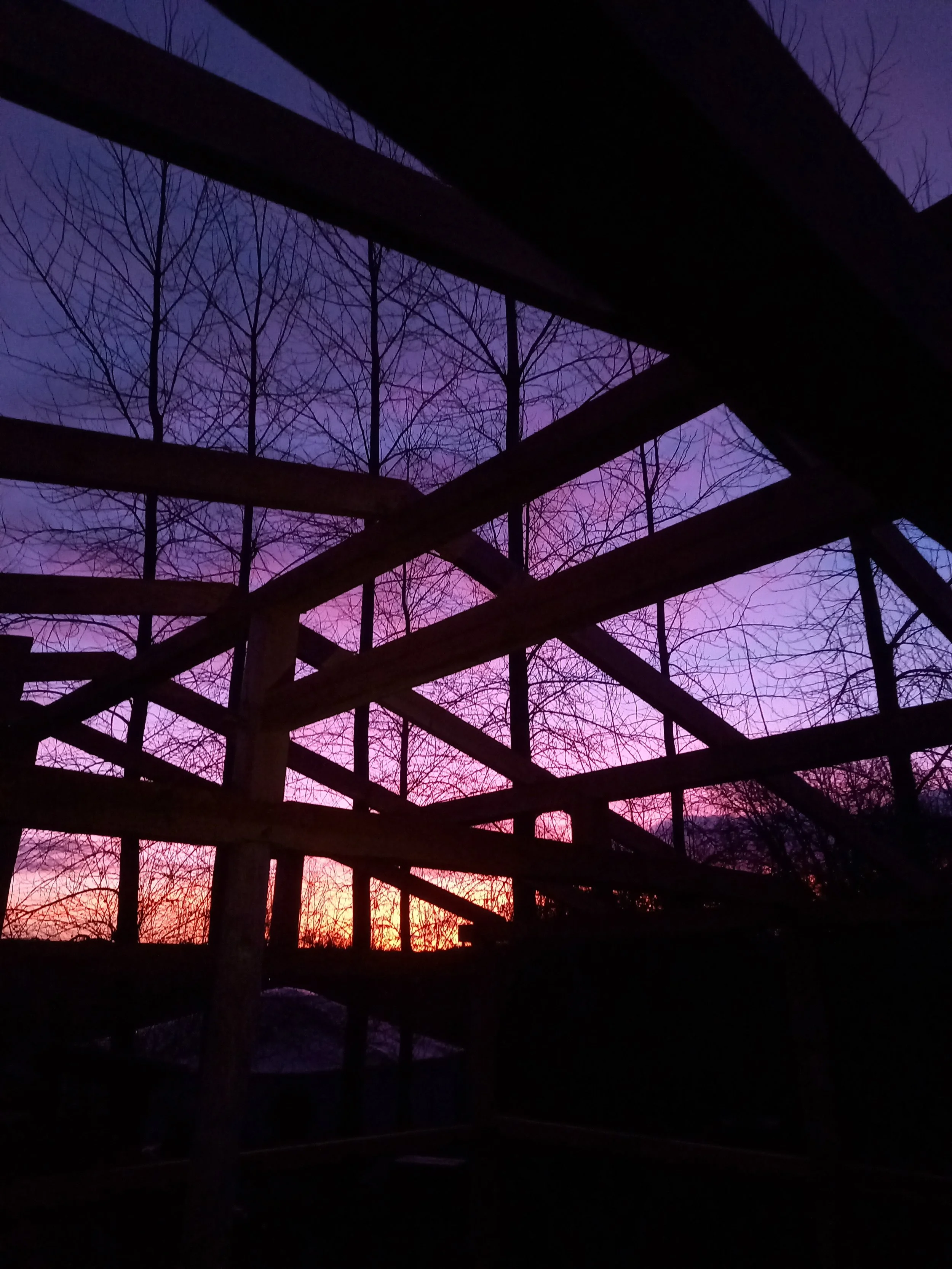 Silhouette of a wooden structure against a colorful sunset sky with leafless trees in the background.