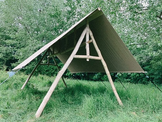 A wooden A-frame shelter with a dark roof supported by three main wooden poles and a cable tie-down in a grassy area surrounded by trees.