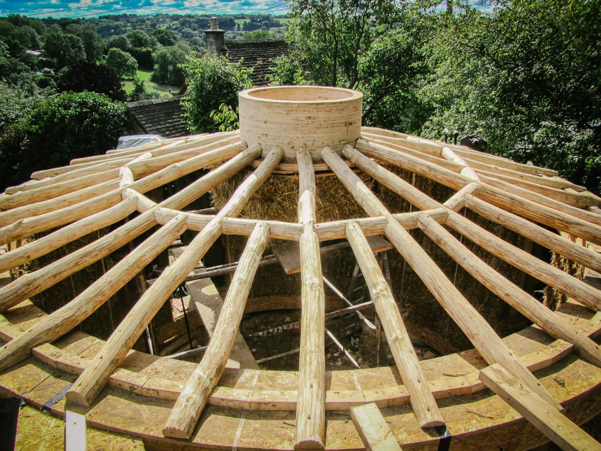 Construction site of a roof with wooden beams arranged in a radial pattern, with a circular wooden structure at the center, outdoors with trees and houses in the background.