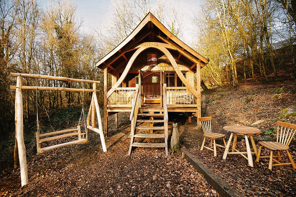 A small wooden treehouse with stairs, a porch, and a swing hanging from a support beam in a wooded area, with a small round table and two chairs nearby.