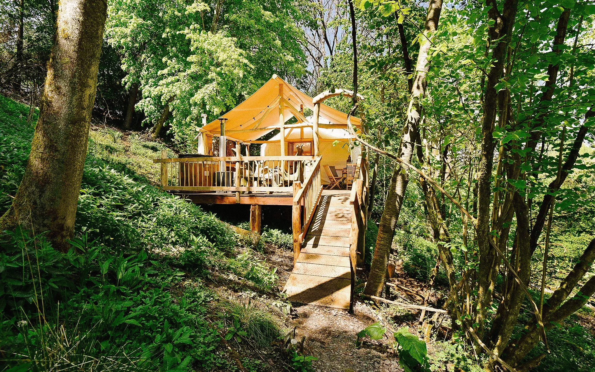 A cozy treehouse with a yellow canopy, situated among green trees and plants in a forest setting. It has a wooden ramp leading up to the entrance and a surrounding deck with outdoor furniture.