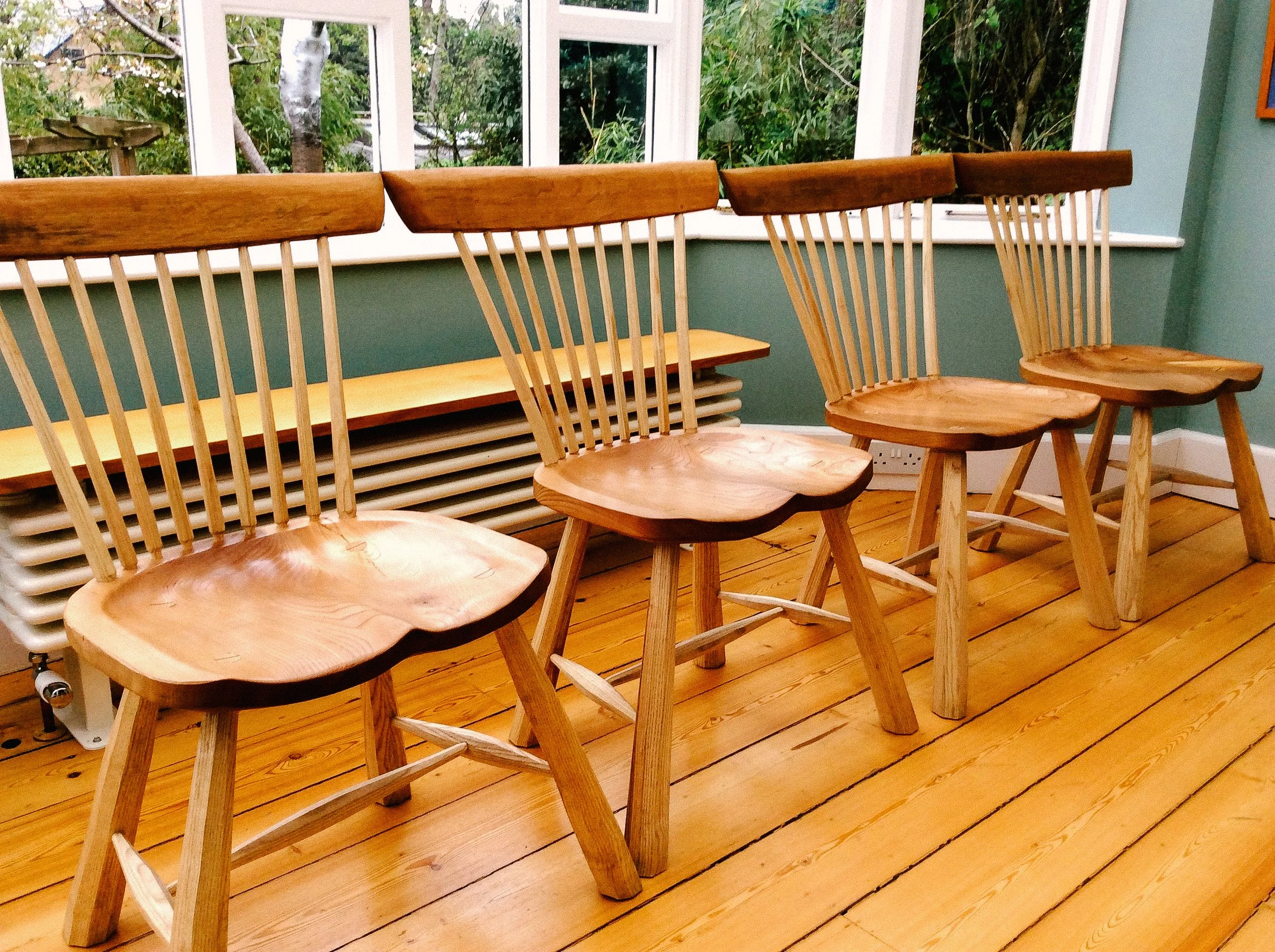 Four wooden chairs with carved seats and high backrests are lined up along a wooden floor in front of a window, with a green wall behind.