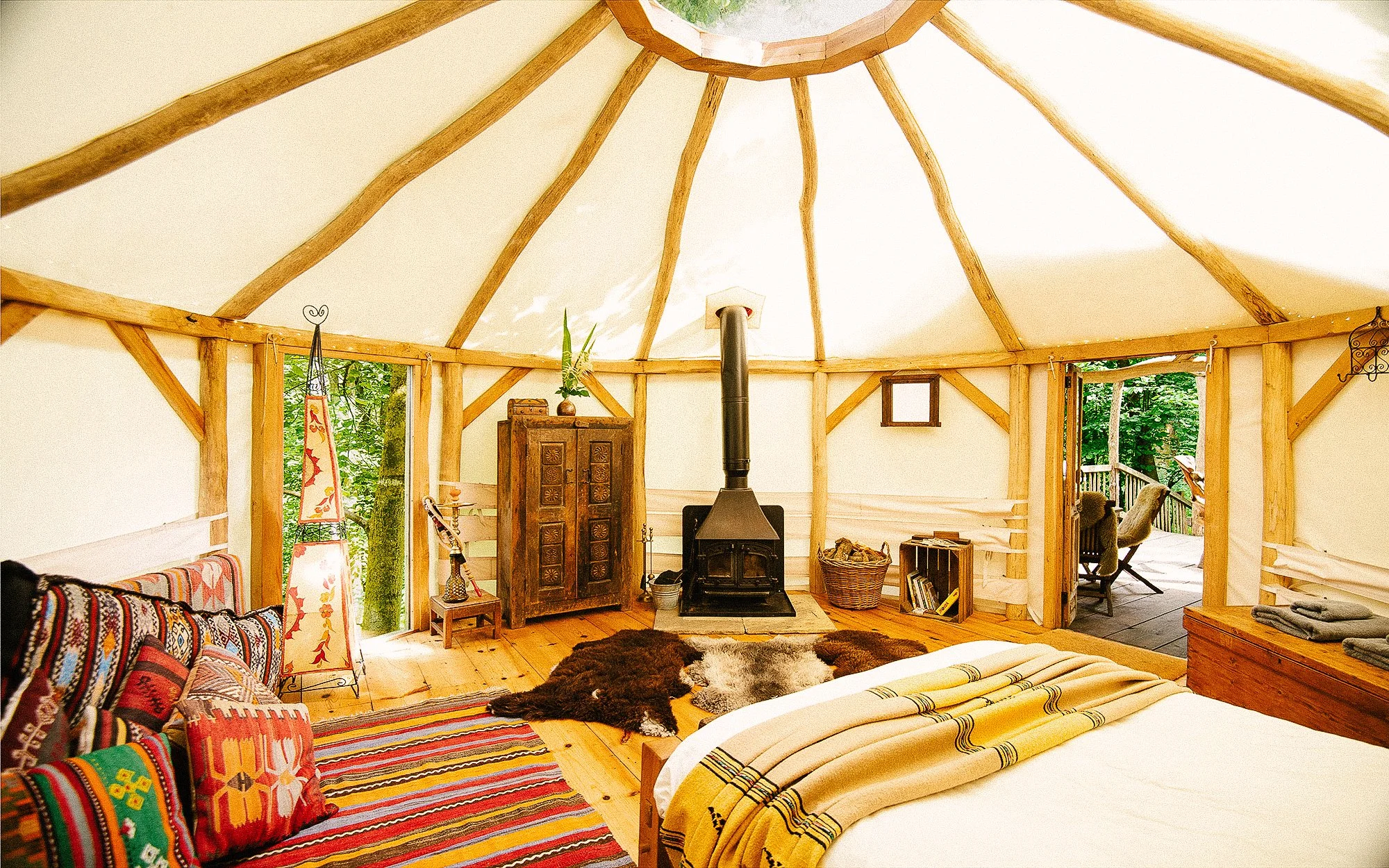 Interior of a cozy, rustic glamping tent with wooden beams, a wood stove, and a wooden deck visible through an open door.