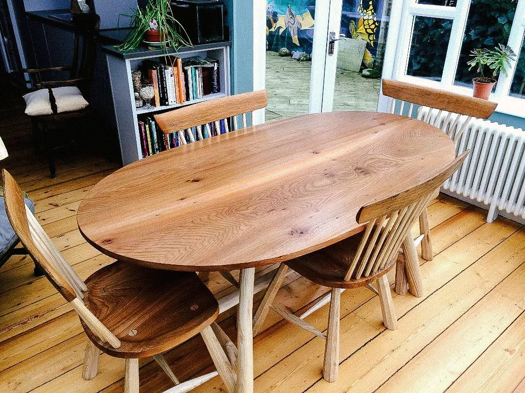 Wooden dining table with four matching wooden chairs in a bright room with hardwood floors, a bookshelf, potted plants, and a sliding glass door leading outside.