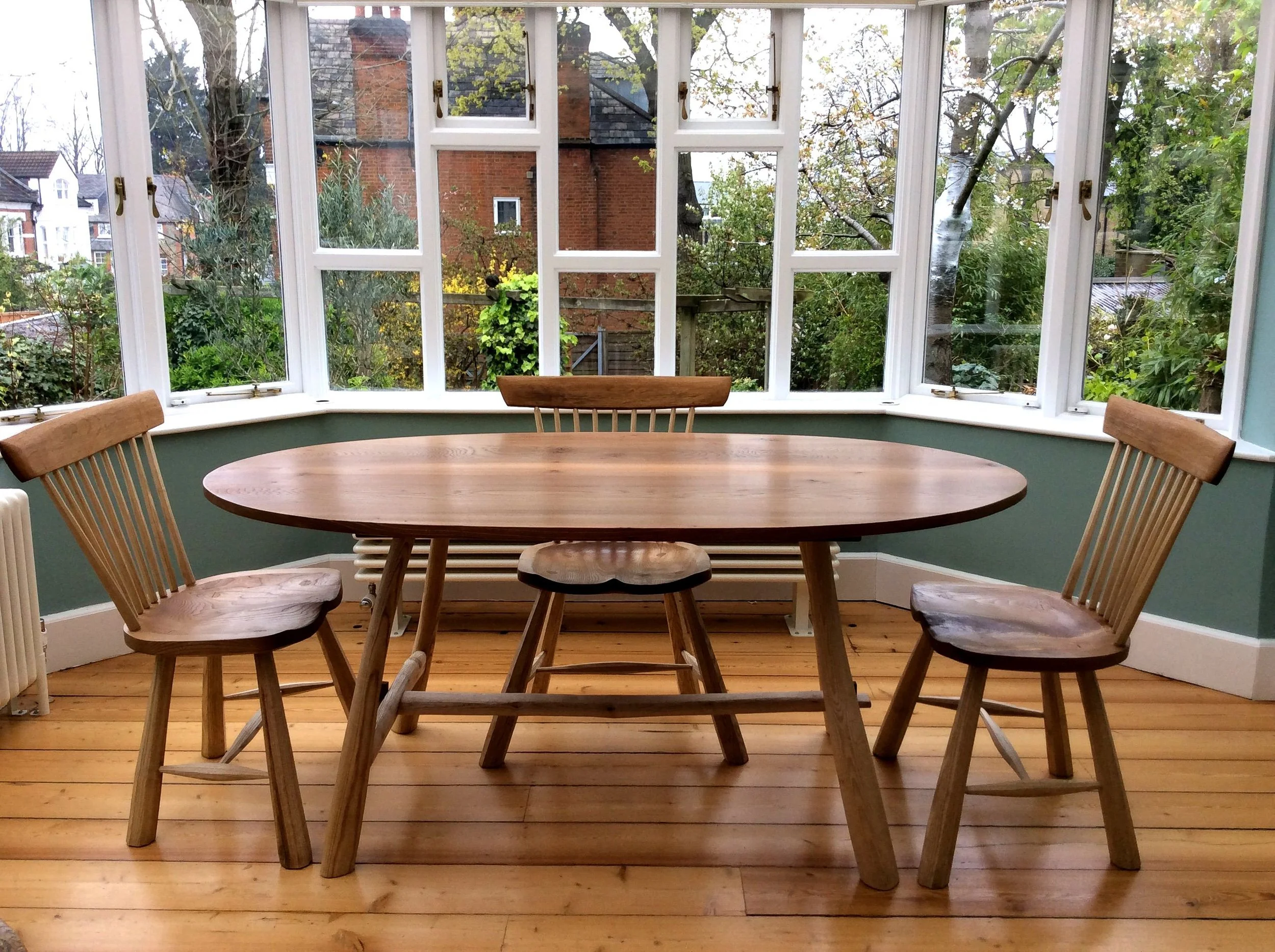 A wooden dining table with four matching wooden chairs in a sunroom with large windows and a view of a garden and neighboring houses.