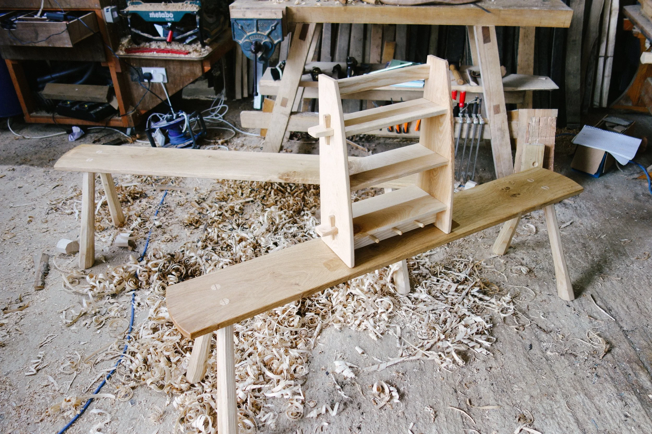A woodworking project in progress in a carpentry workshop, featuring a partially assembled wooden bench with a raised shelf on one end, surrounded by wood shavings and woodworking tools.