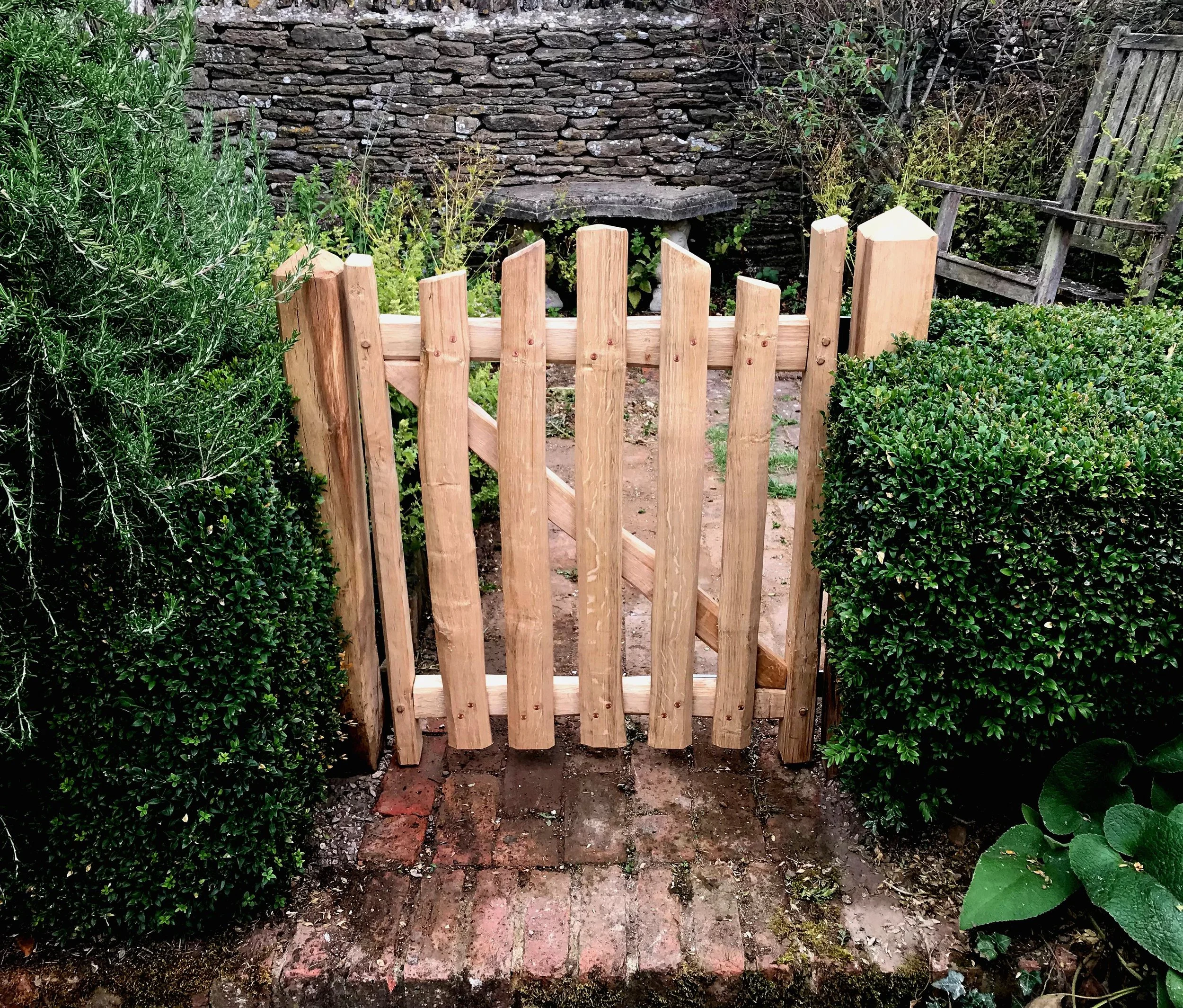 Small wooden garden gate flanked by neatly trimmed green bushes, with a stone and brick pathway leading to it and a stone wall background.