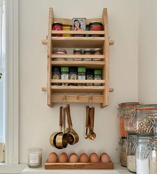 Wooden spice rack mounted on a wall holding jars of spices, with hanging measuring cups and spoons below, and a row of eggs on a shelf underneath.