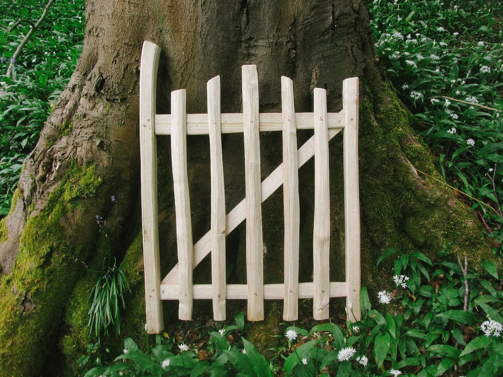 A small, white wooden fence placed against a large tree trunk in a lush green forest, surrounded by moss and white flowers.