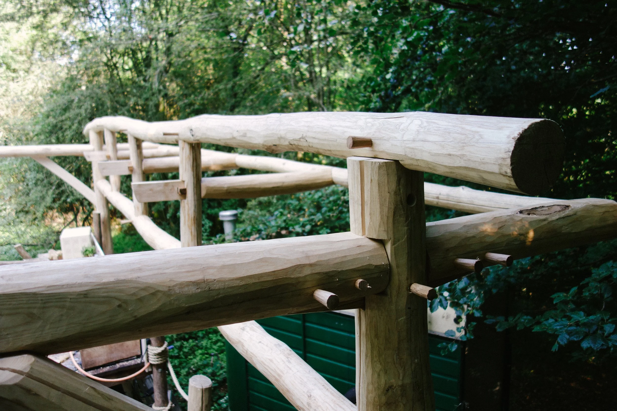 Close-up view of a wooden railing structure in a natural outdoor setting, with trees and greenery in the background.