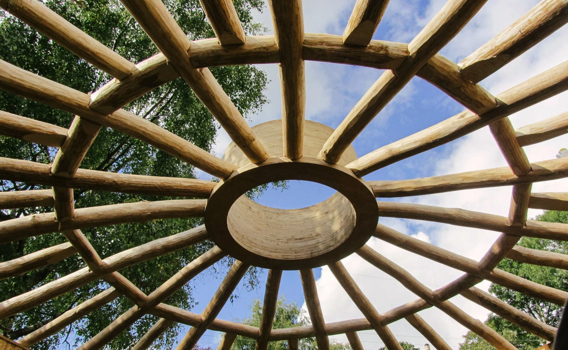 View looking up through a wooden circular shelter with radiating beams, blue sky, and green trees visible in the background.