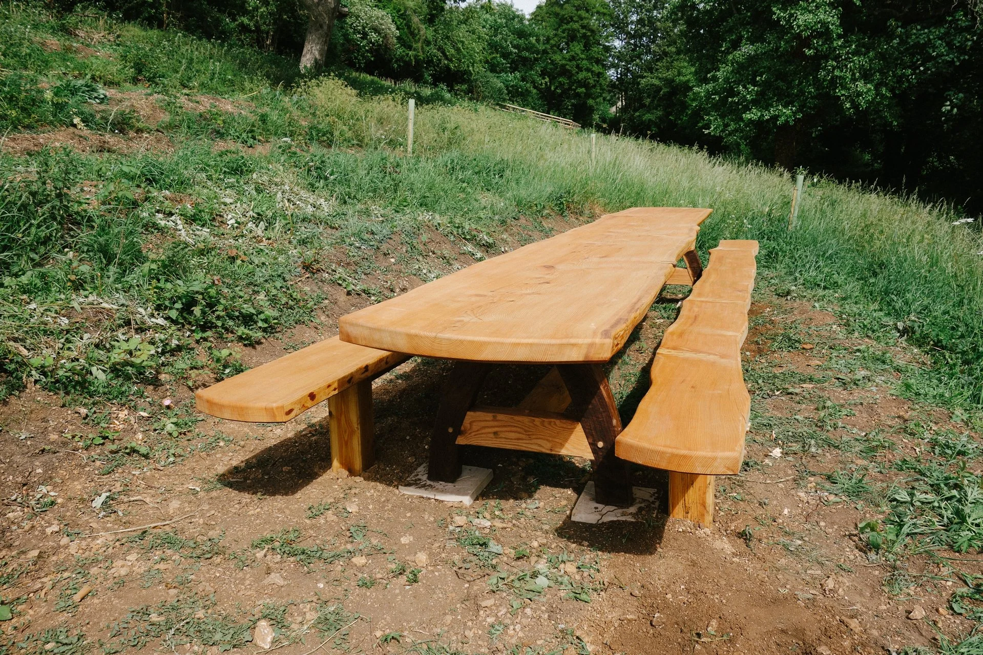 A rustic outdoor wooden picnic table with two long benches on either side, situated on a patch of dirt and grass with greenery and trees in the background.