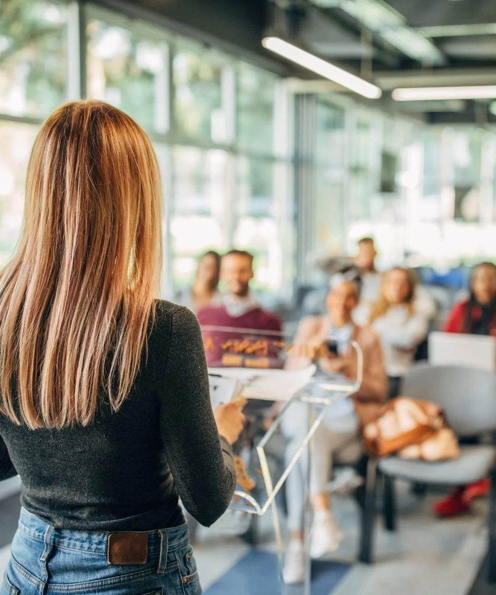 A woman with straight light brown hair giving a presentation or speech to a group of people seated in a bright, modern room with large windows.